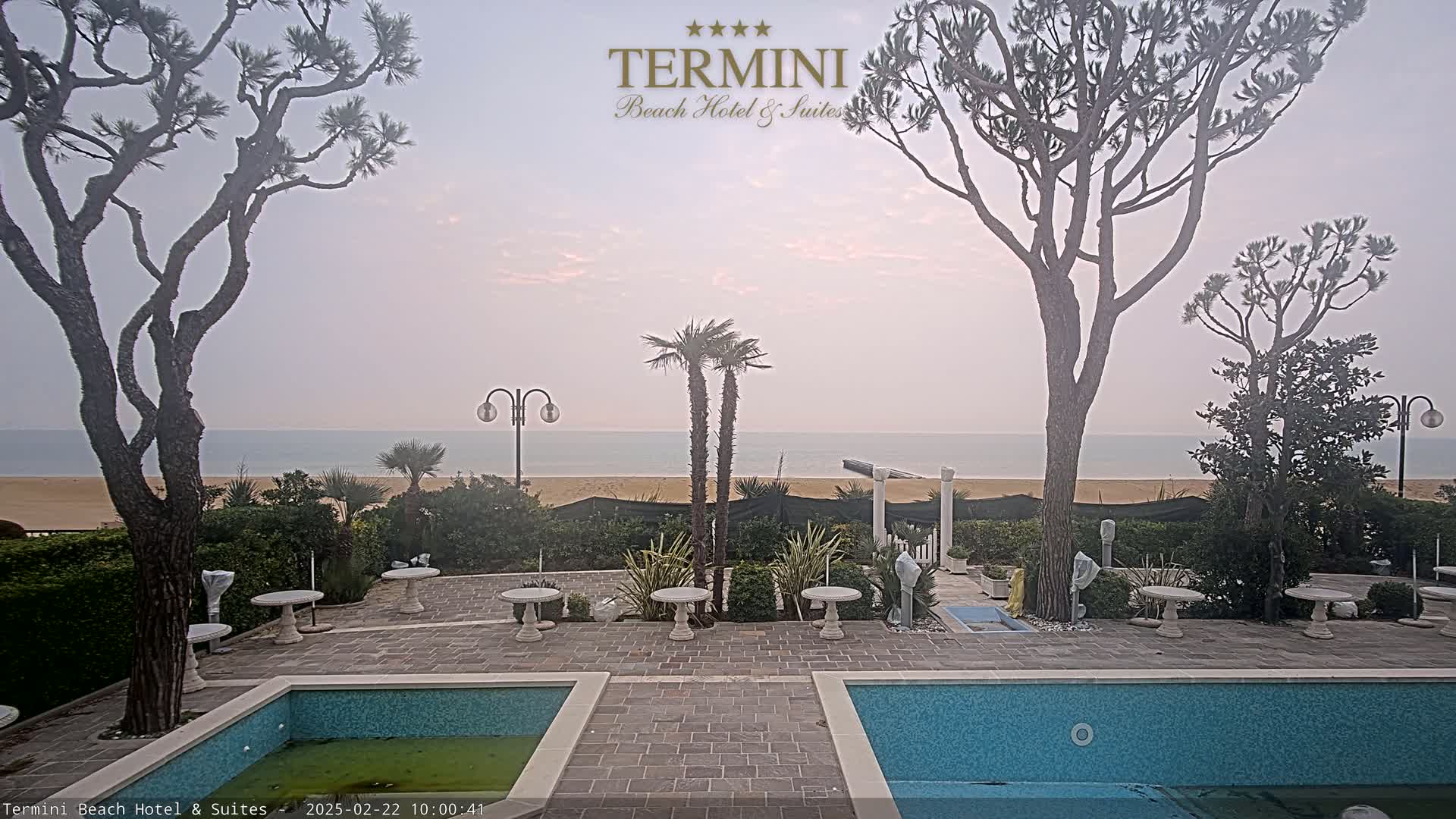 A hazy sunrise illuminates a courtyard with two swimming pools, several small tables, and various trees, overlooking a calm ocean and sandy beach.