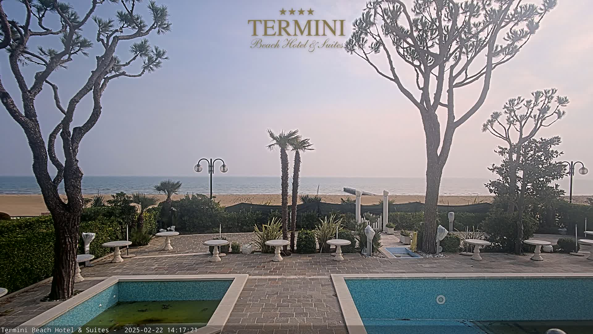 A courtyard with two pools overlooks a calm ocean beach under a hazy sky.