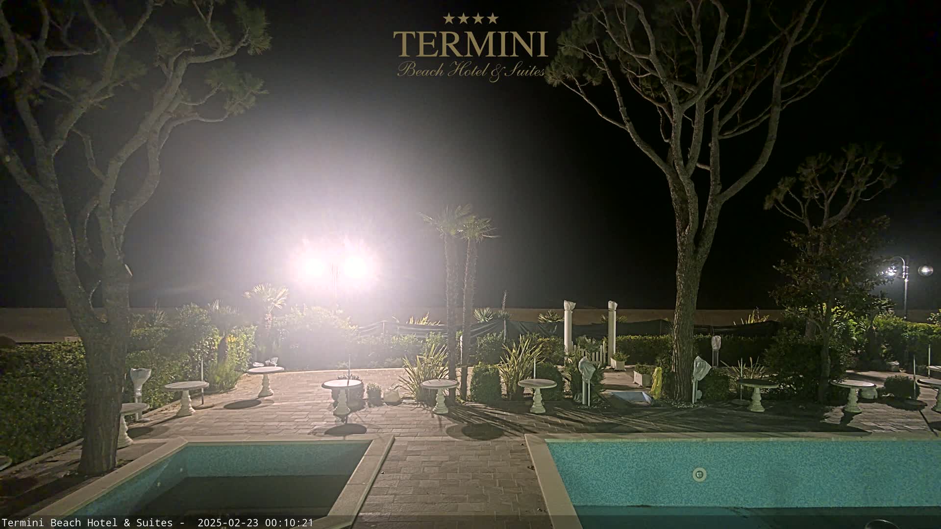 A nighttime view of a patio area with several small tables and chairs surrounding a swimming pool, illuminated by bright lights under a dark sky.