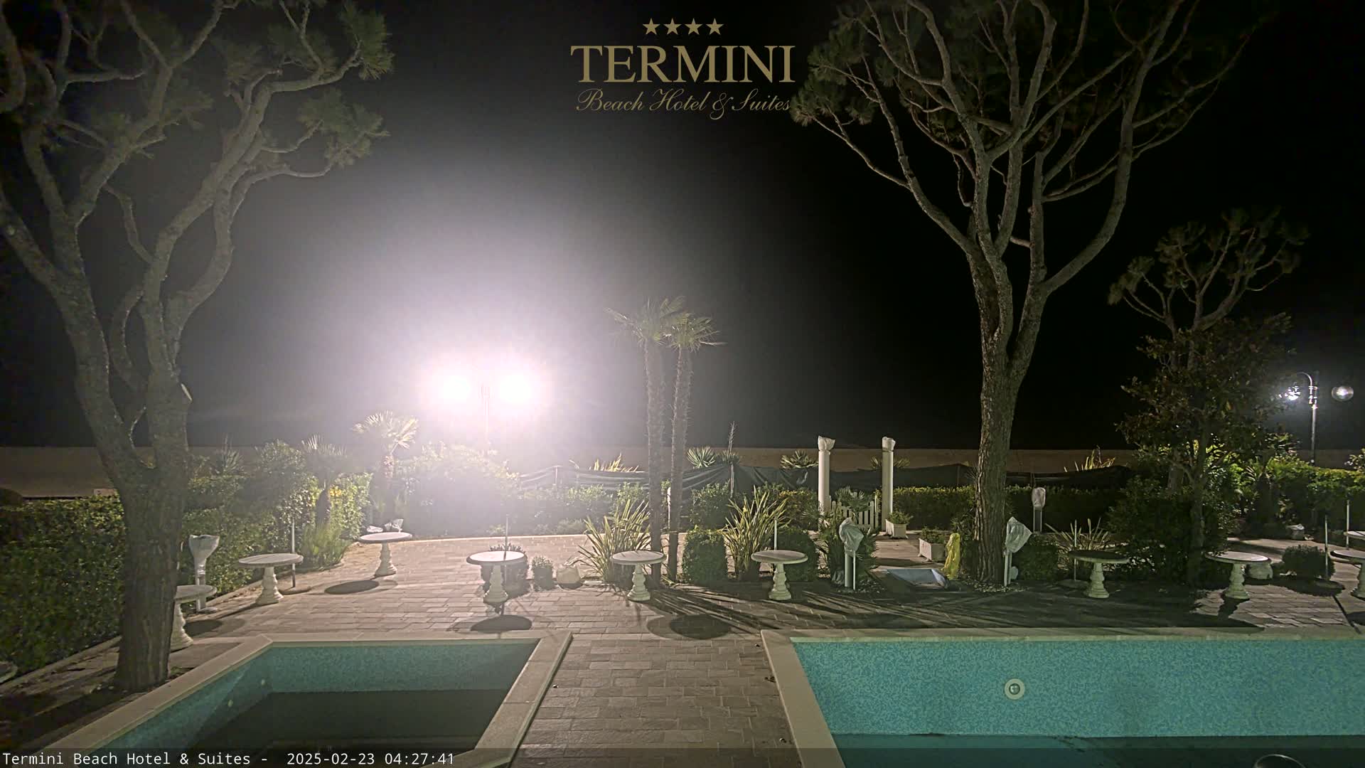 At night, a brightly lit patio area with two swimming pools is surrounded by trees and shrubs.