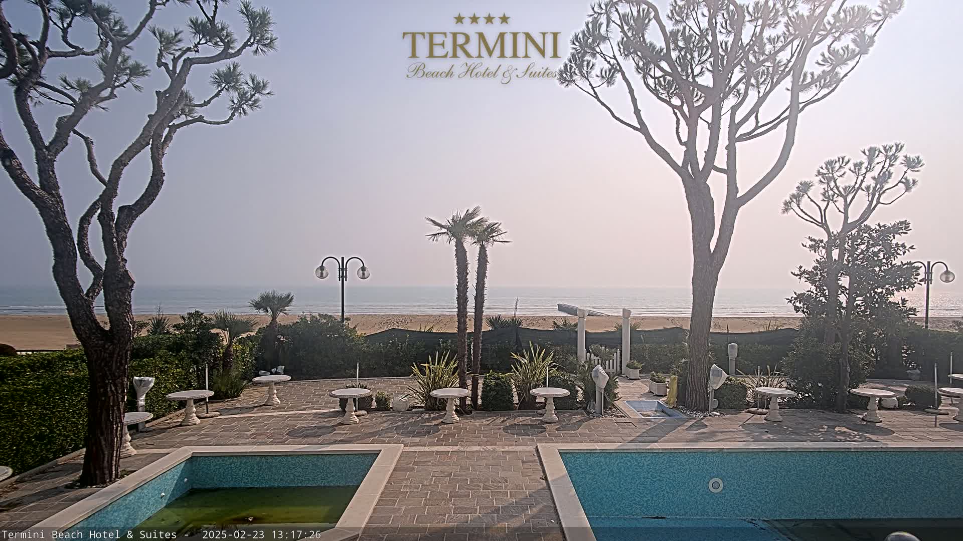 A patio with two swimming pools overlooks a calm ocean beach on a hazy day.