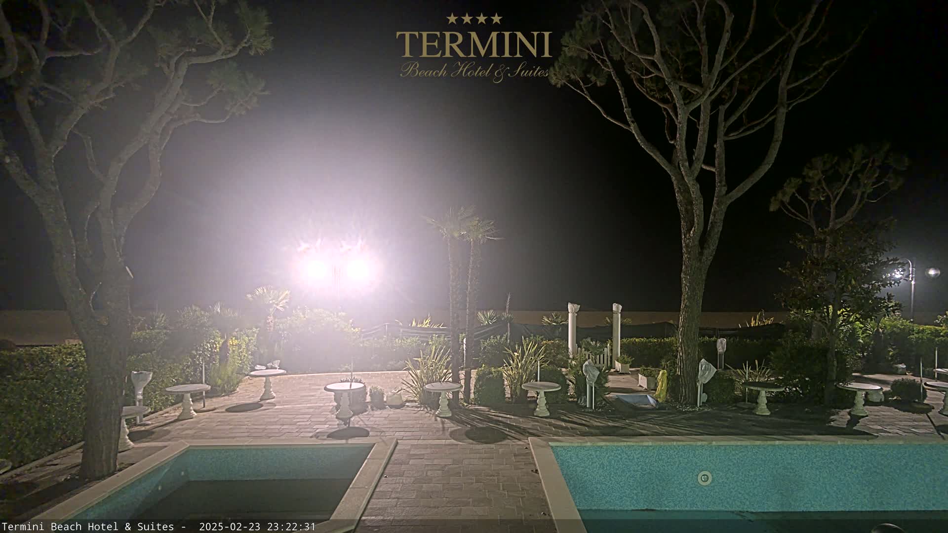 At night, under hazy lighting conditions, an outdoor area with two swimming pools, several small tables, and trees is visible.