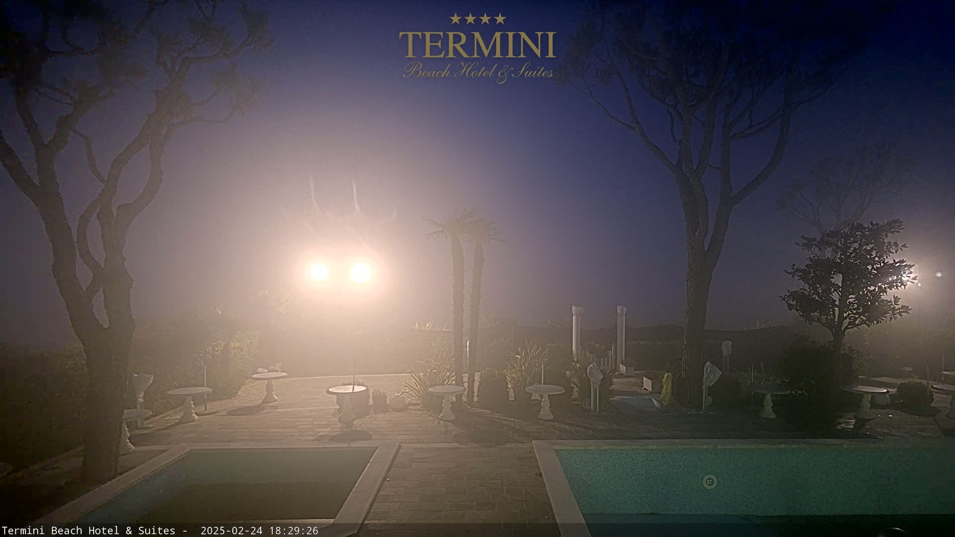 A hazy, twilight scene shows an outdoor pool area with two pools, surrounding landscaping, and several small tables and chairs, illuminated by bright lights.