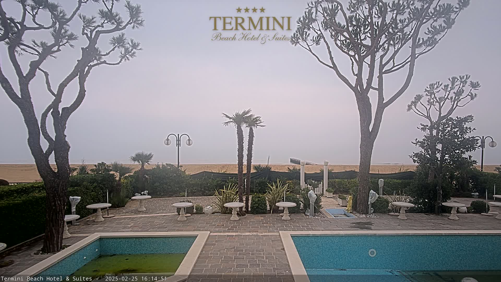 A hazy, overcast day reveals a courtyard with two pools, stone tables, and several trees, overlooking a sandy beach.