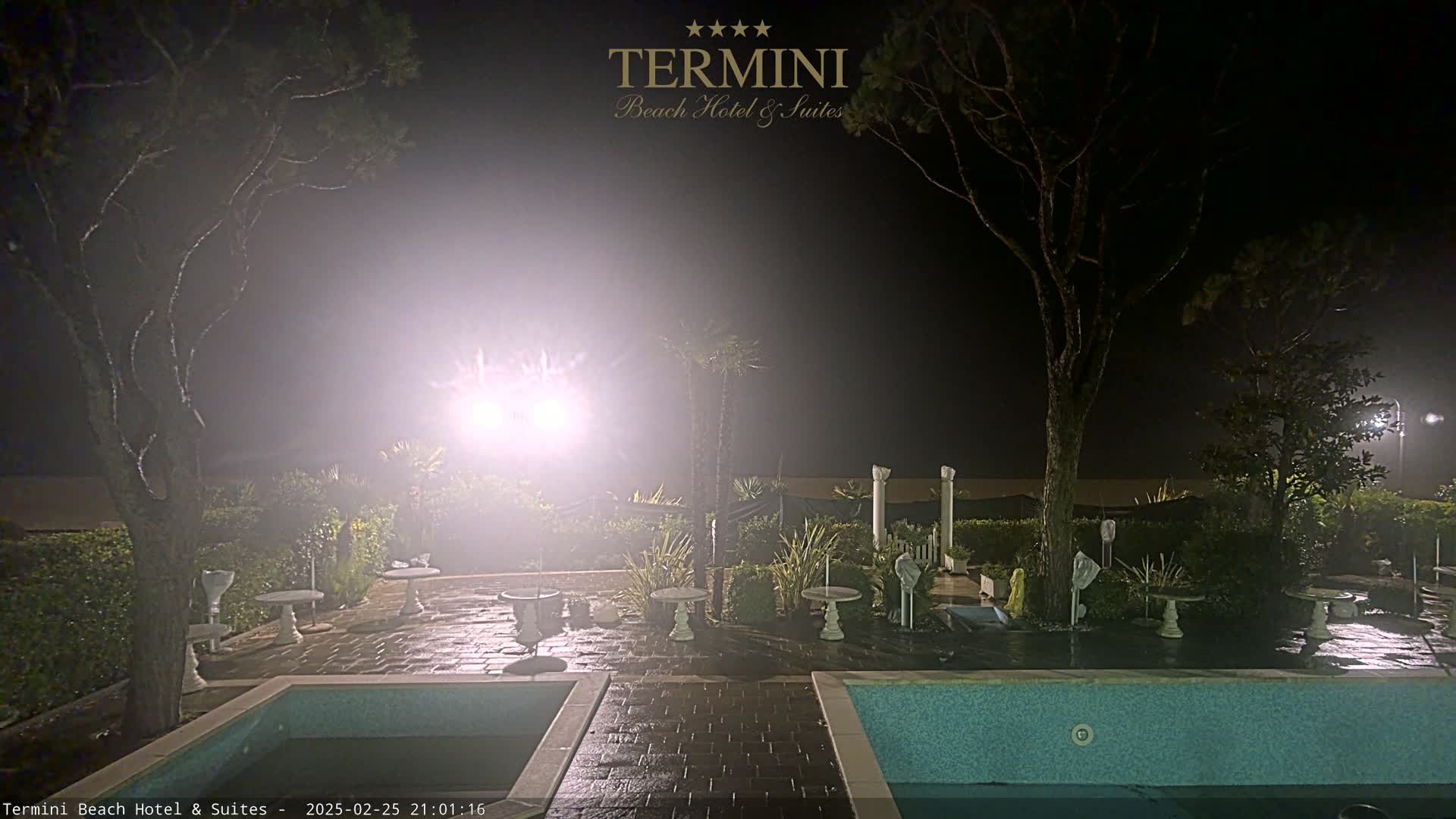 A hazy nighttime scene shows a patio area with two pools, small tables, and trees, illuminated by bright lights.