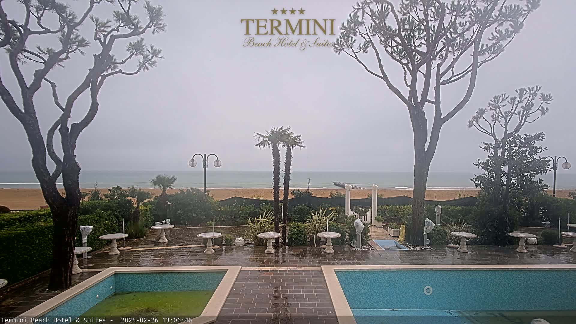 A rain-swept view of a resort area with two swimming pools, several small tables and chairs, and palm and pine trees overlooking a sandy beach and ocean under an overcast sky.