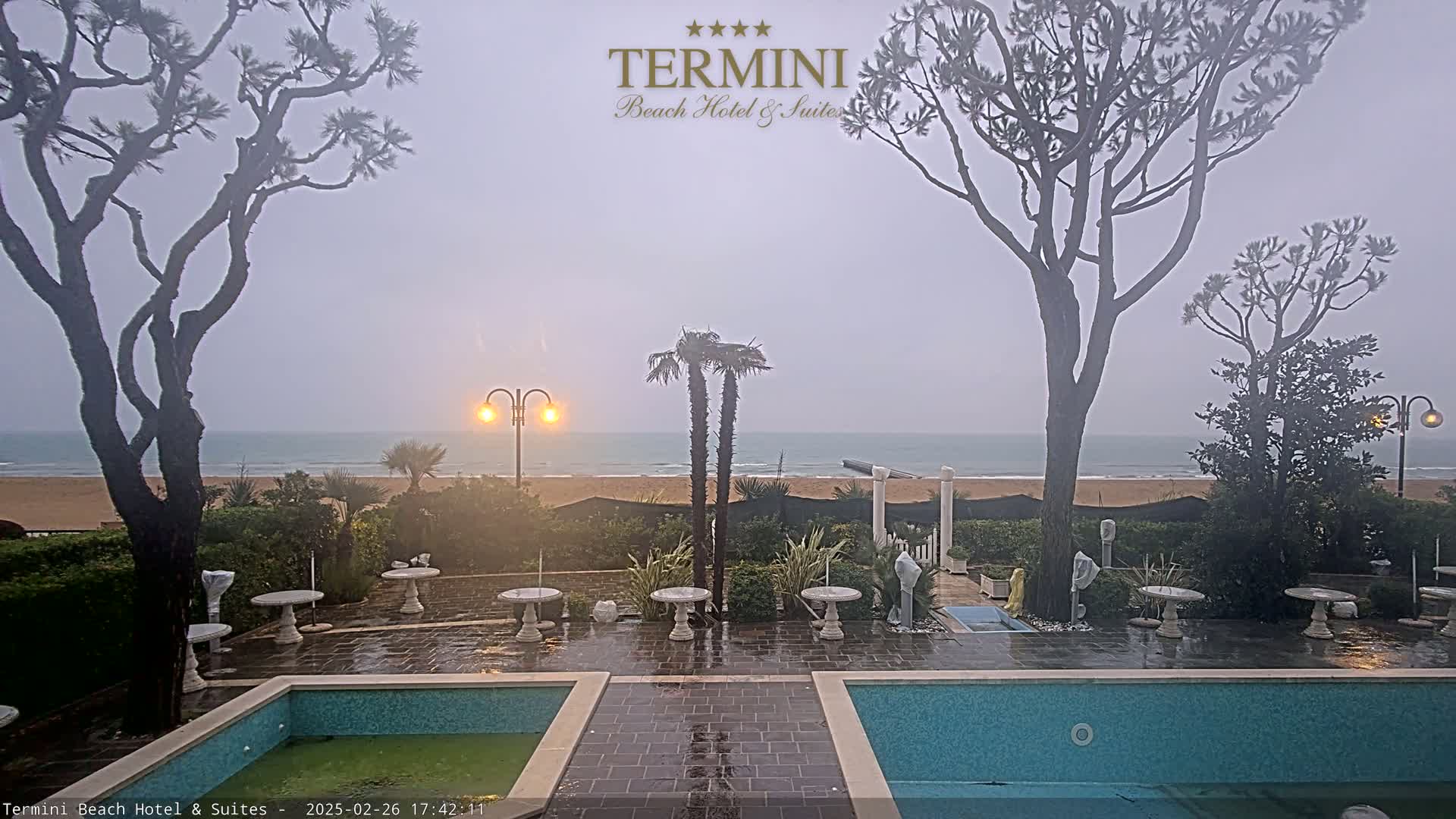 A rain-swept outdoor patio scene shows a calm ocean in the distance, two pools, several small tables, and tall trees surrounding the area.