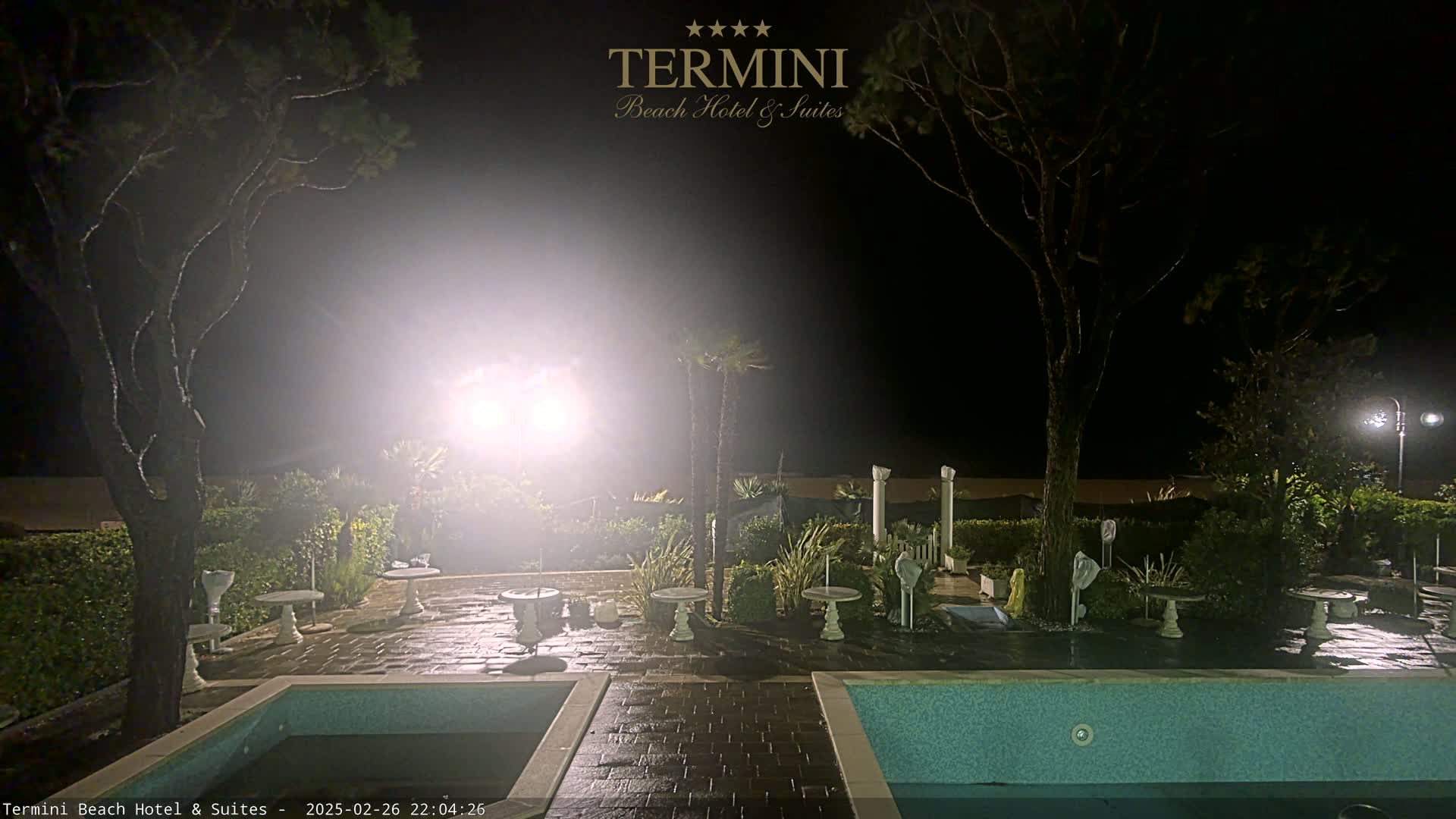 A nighttime view of a patio area with two pools, several small tables, and surrounding landscaping, illuminated by bright lights in hazy conditions.