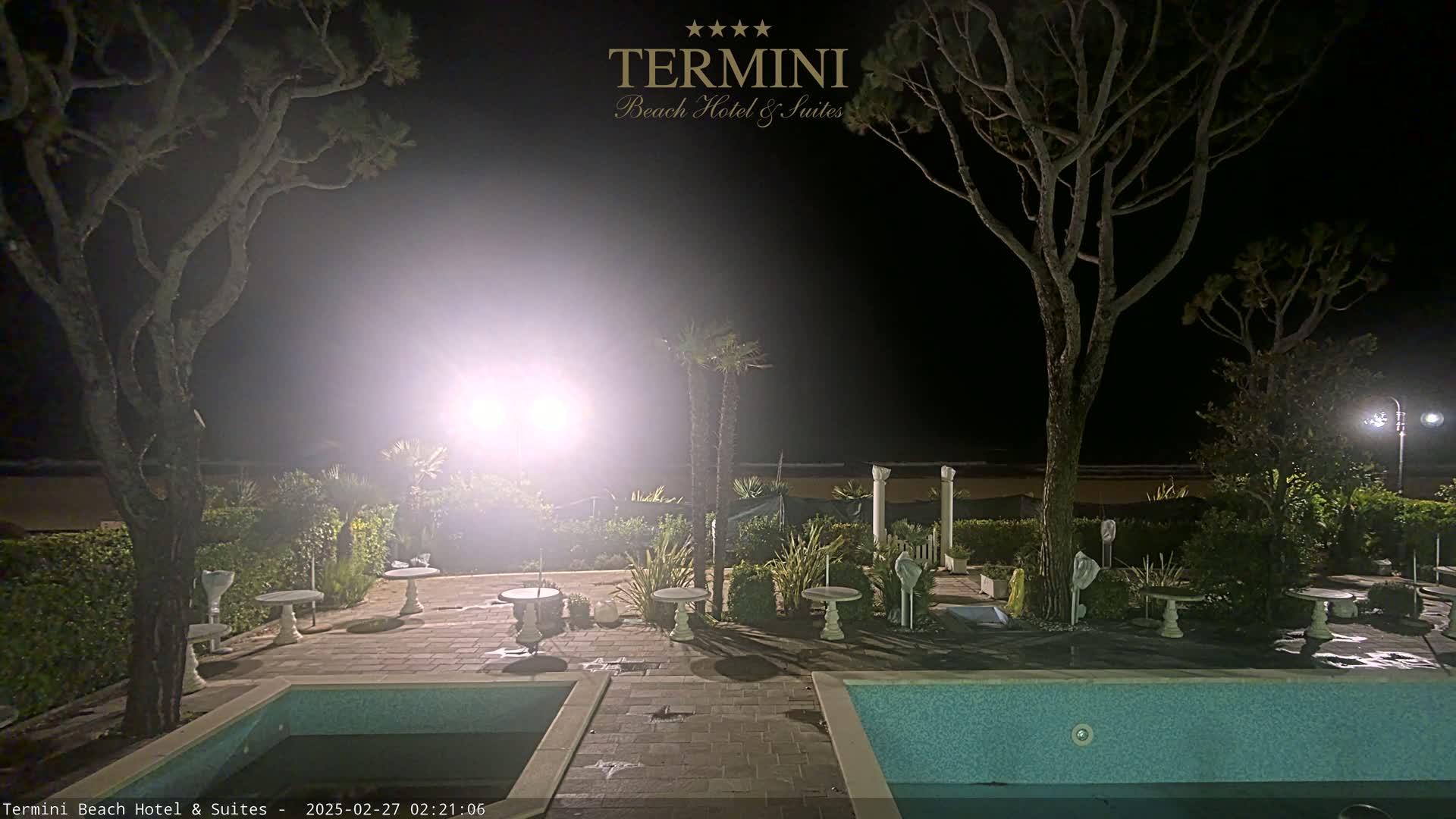 At night, a brightly lit patio area with several small tables and chairs surrounds a swimming pool, situated between two tall, dark trees near a beach under hazy conditions.