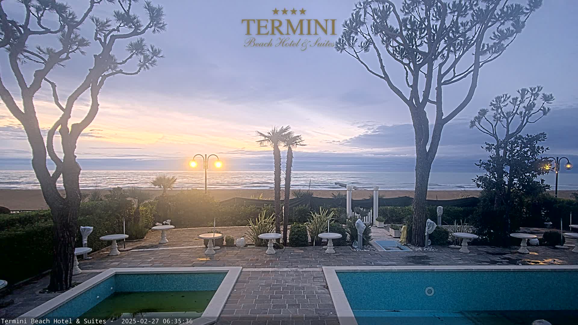 A tranquil seaside patio scene at sunrise features two swimming pools, outdoor seating, and palm trees, all overlooking a calm ocean under a soft, pastel sky.