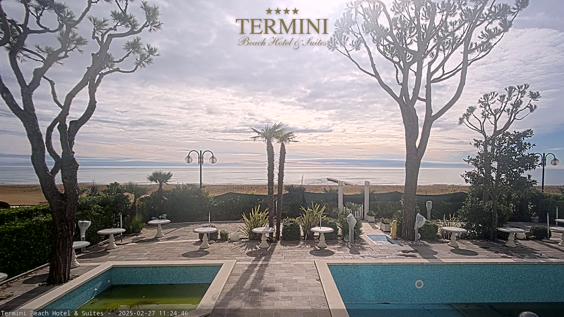 A courtyard with two swimming pools overlooks a calm ocean beach under a mostly cloudy sky.