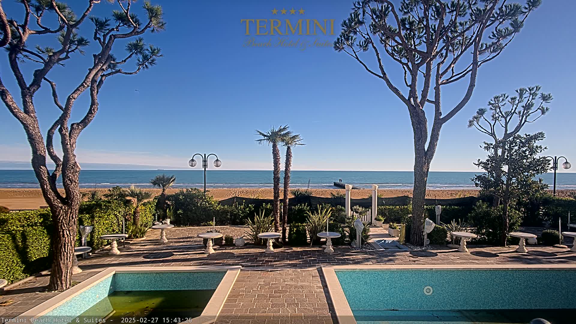 A patio area with two small pools overlooks a sandy beach and calm ocean under a clear blue sky.