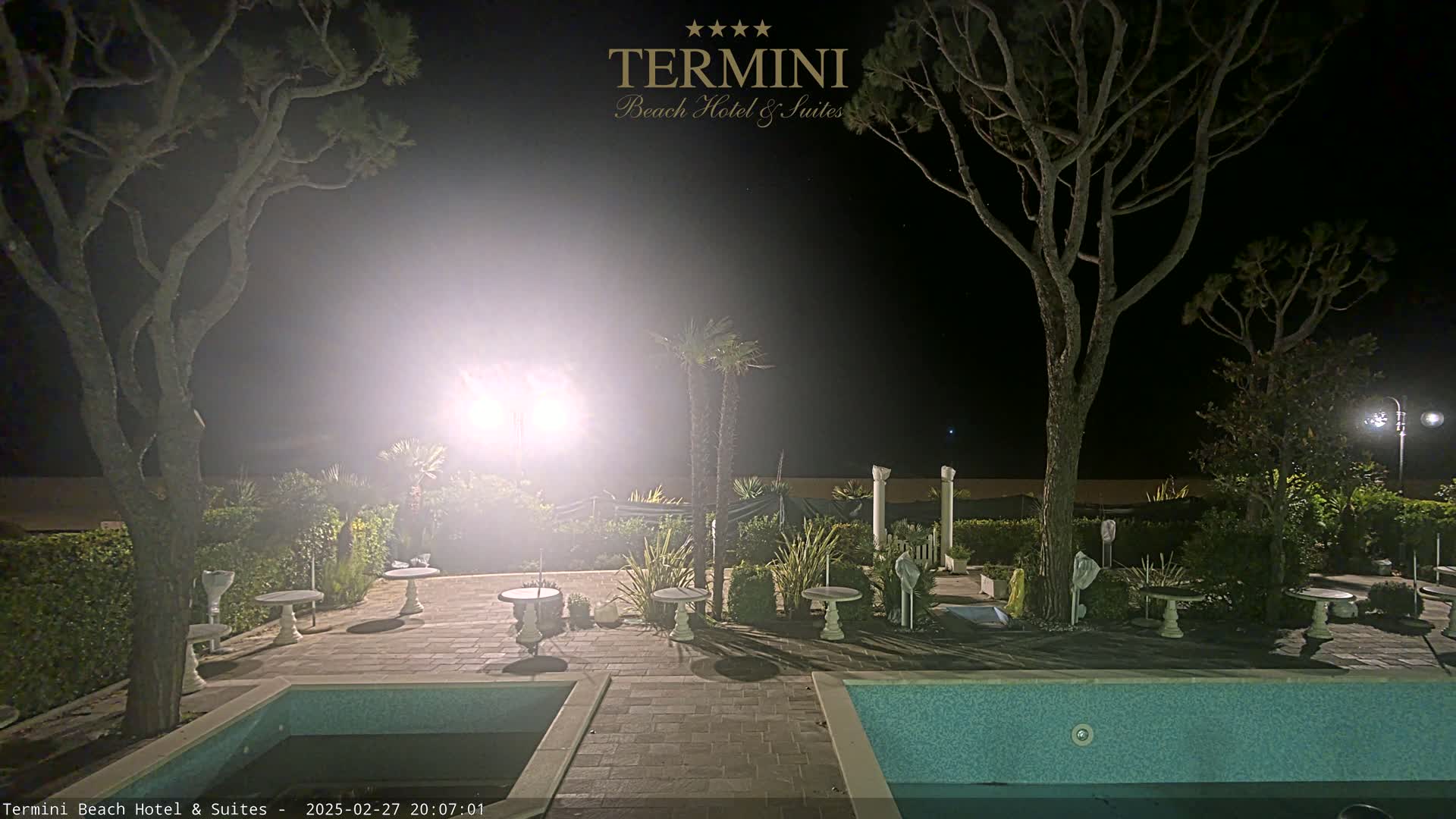 A nighttime view of a patio area with a pool, surrounded by trees and small tables, illuminated by bright lights.
