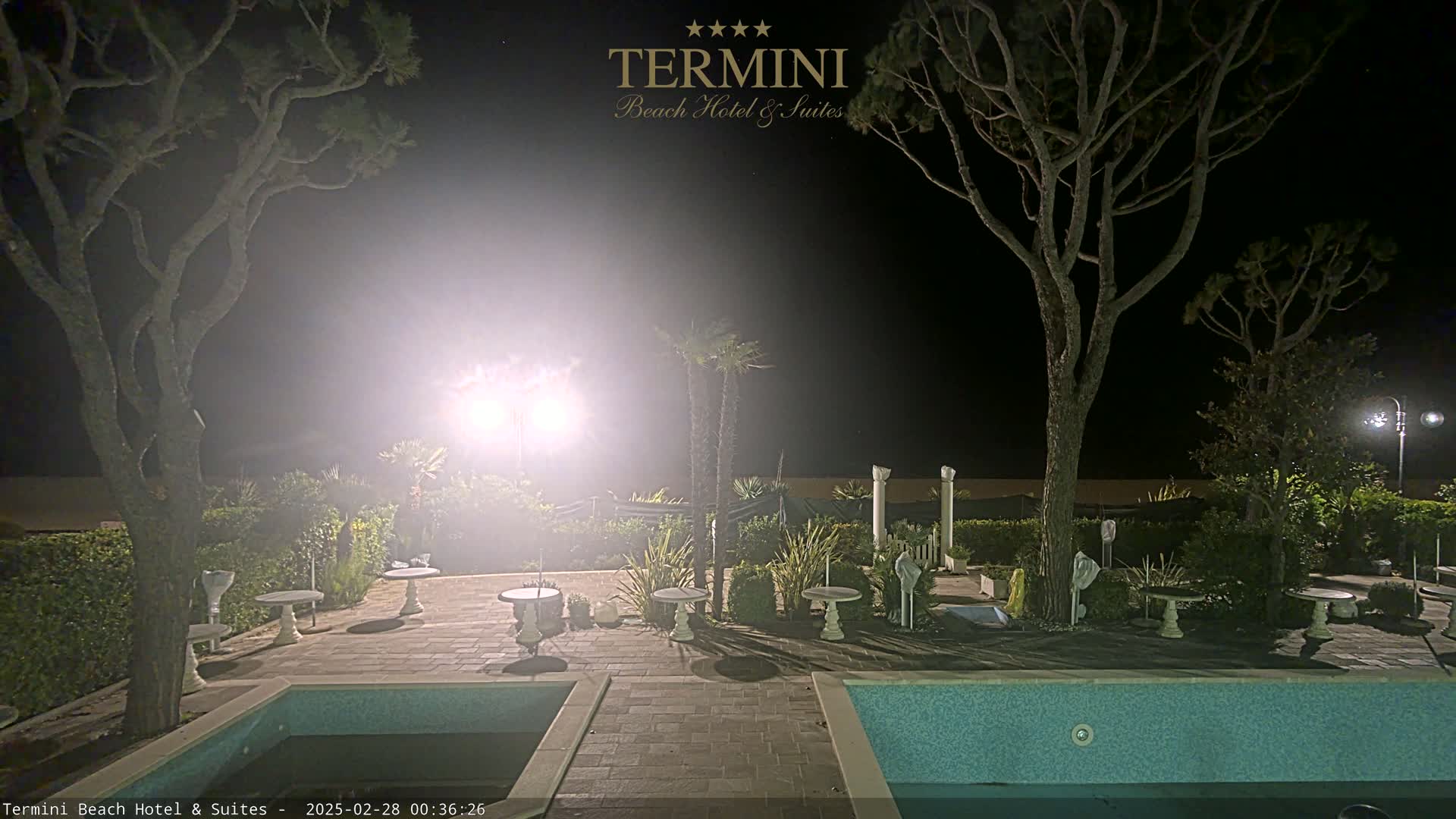 At night, a brightly lit patio area with small tables and a swimming pool is surrounded by trees and hedges.
