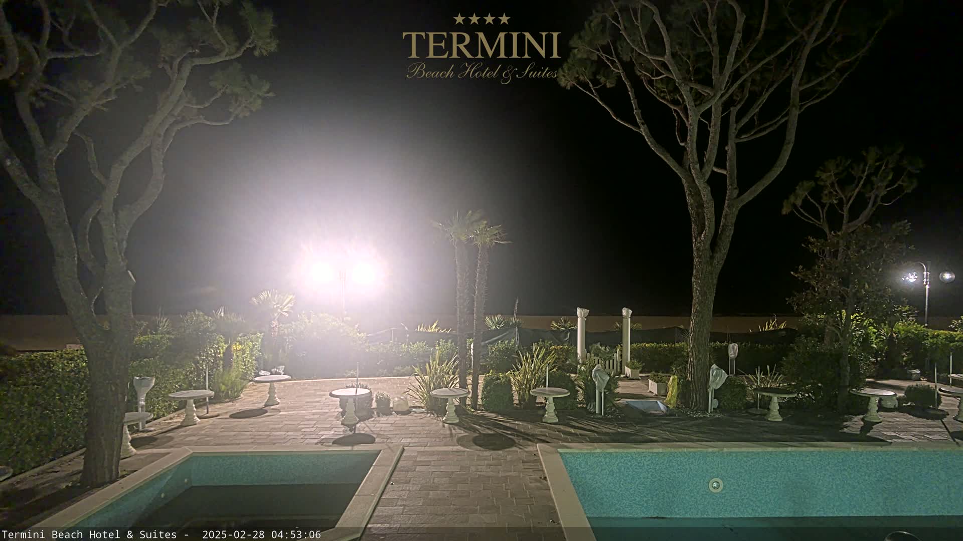 A nighttime view of a hotel pool area with two trees flanking the scene, illuminated by bright lights, and small tables with chairs arranged around it.