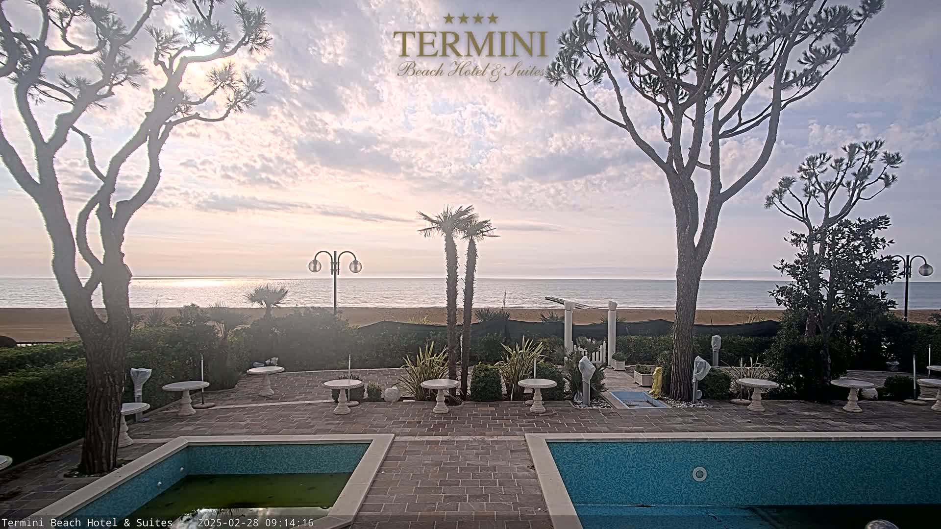 A tranquil outdoor patio scene overlooks a calm ocean under a partly cloudy sky, featuring two swimming pools, several potted plants, and tall trees flanking the view.