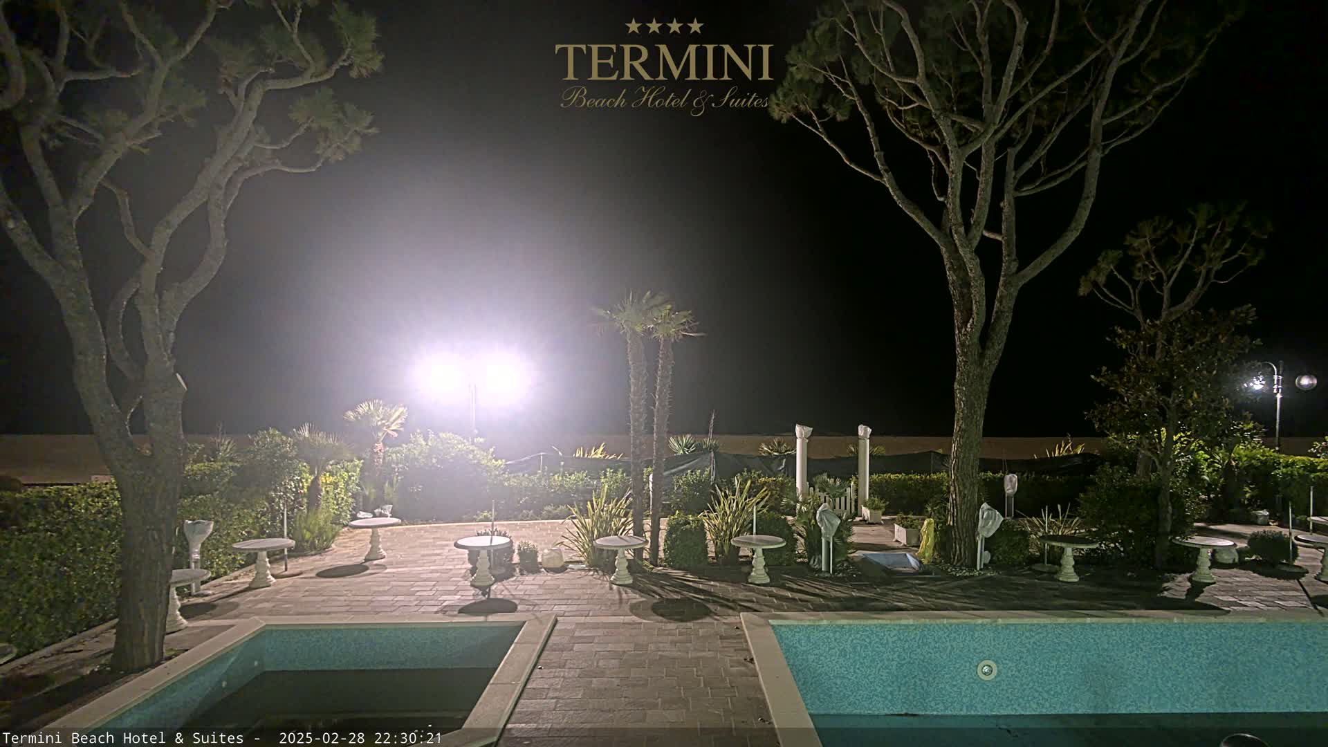 At night, a swimming pool and patio furniture sit under bright lights next to trees and hedges.