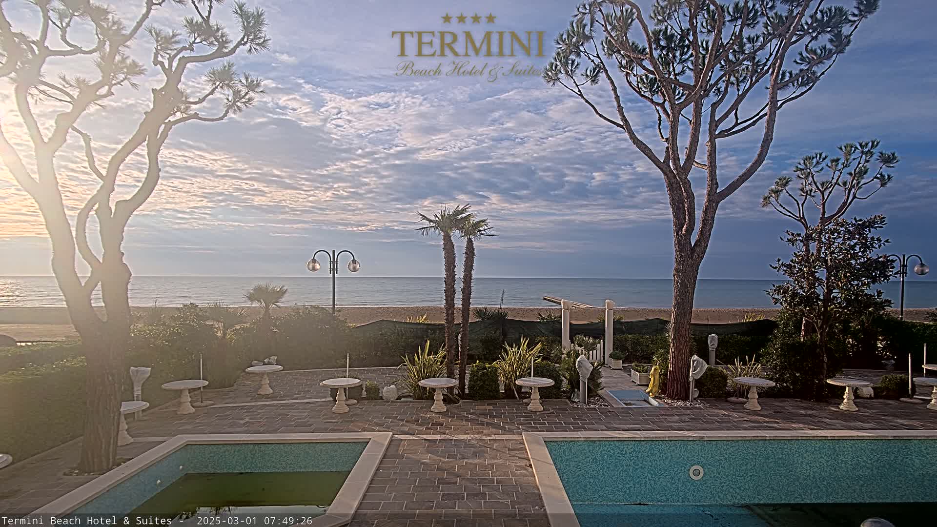 A courtyard with two pools overlooks a calm ocean under a partly cloudy sky.