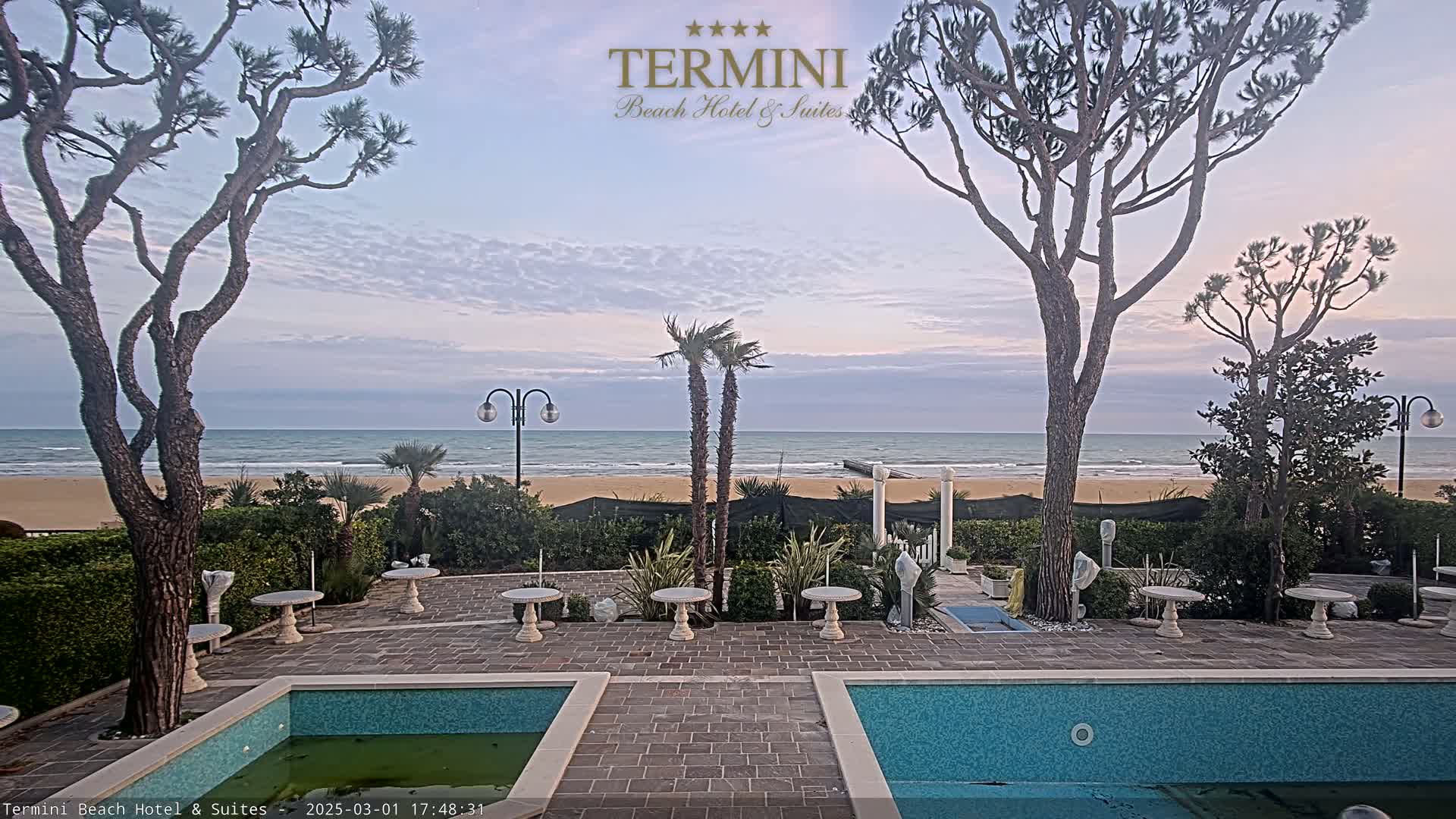 A calm ocean is visible beyond a patio with two pools, several small tables, and trees under a partly cloudy sky.