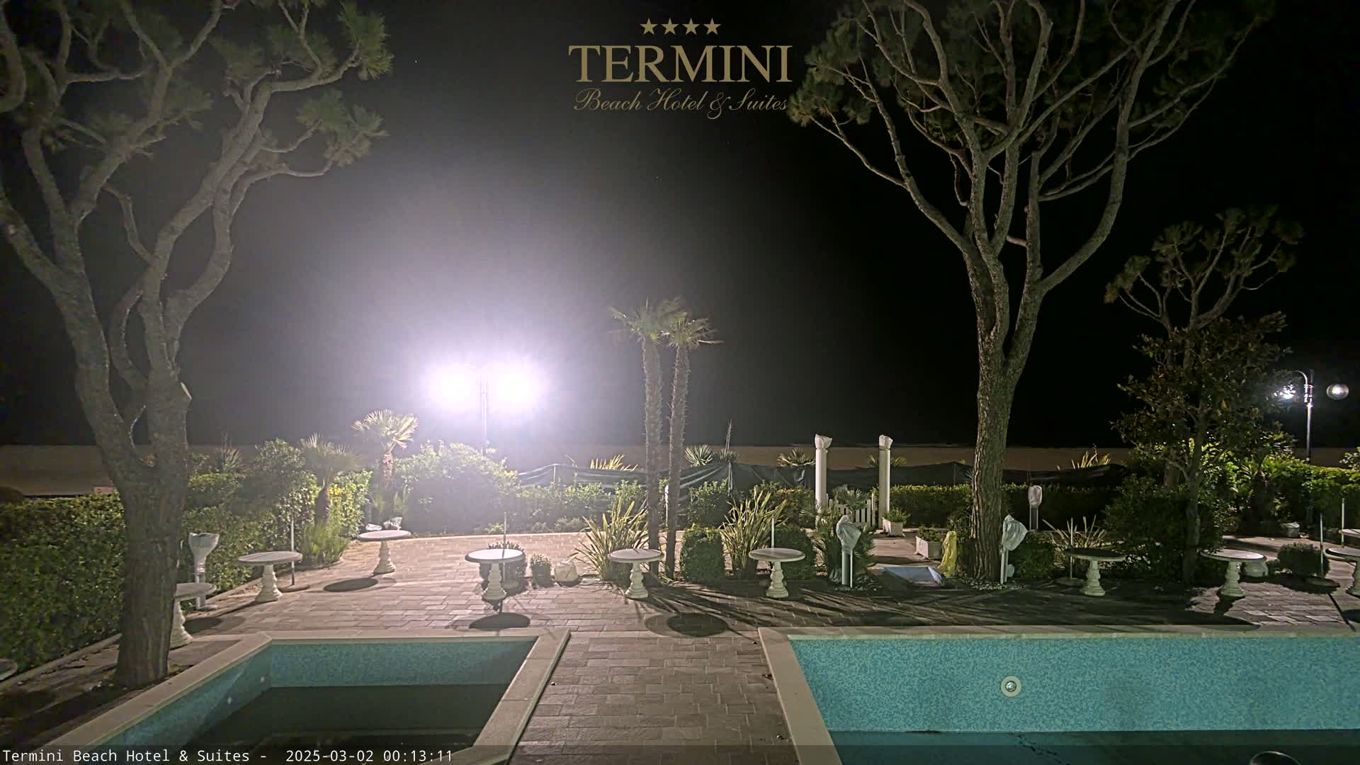 A nighttime view of a patio area with two swimming pools, several small tables, and trees, illuminated by bright lights.