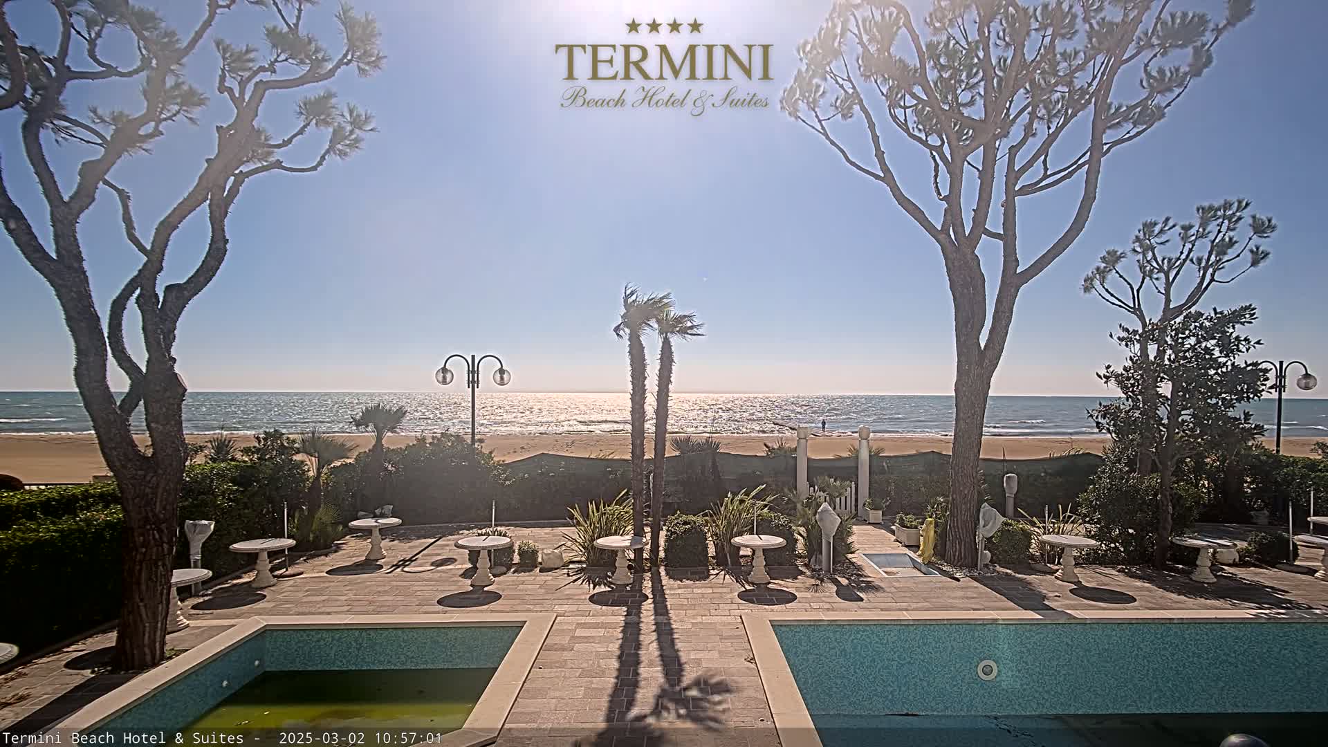 A sunny day at a beachfront hotel reveals a patio with two pools, small tables, and landscaping, overlooking a calm ocean.