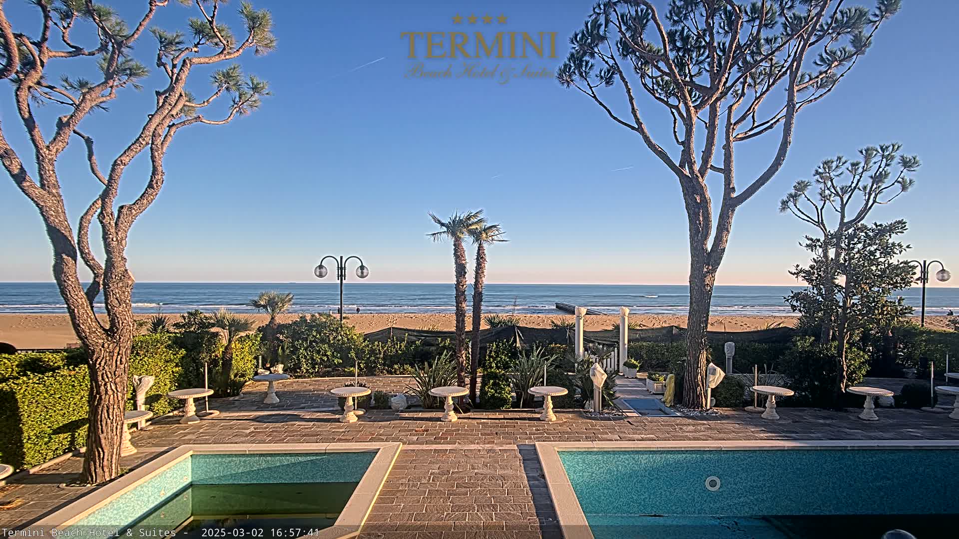 A courtyard with two swimming pools overlooks a sandy beach and ocean under a clear blue sky.