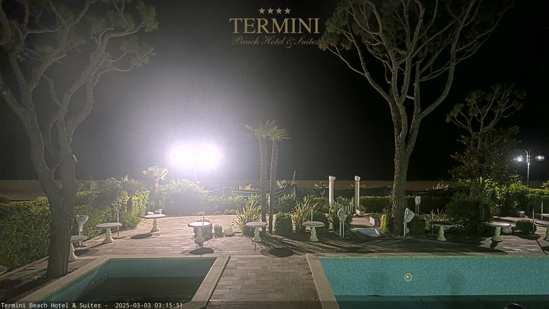 A nighttime view of a resort area with a swimming pool, patio furniture, and palm trees under bright artificial lighting.