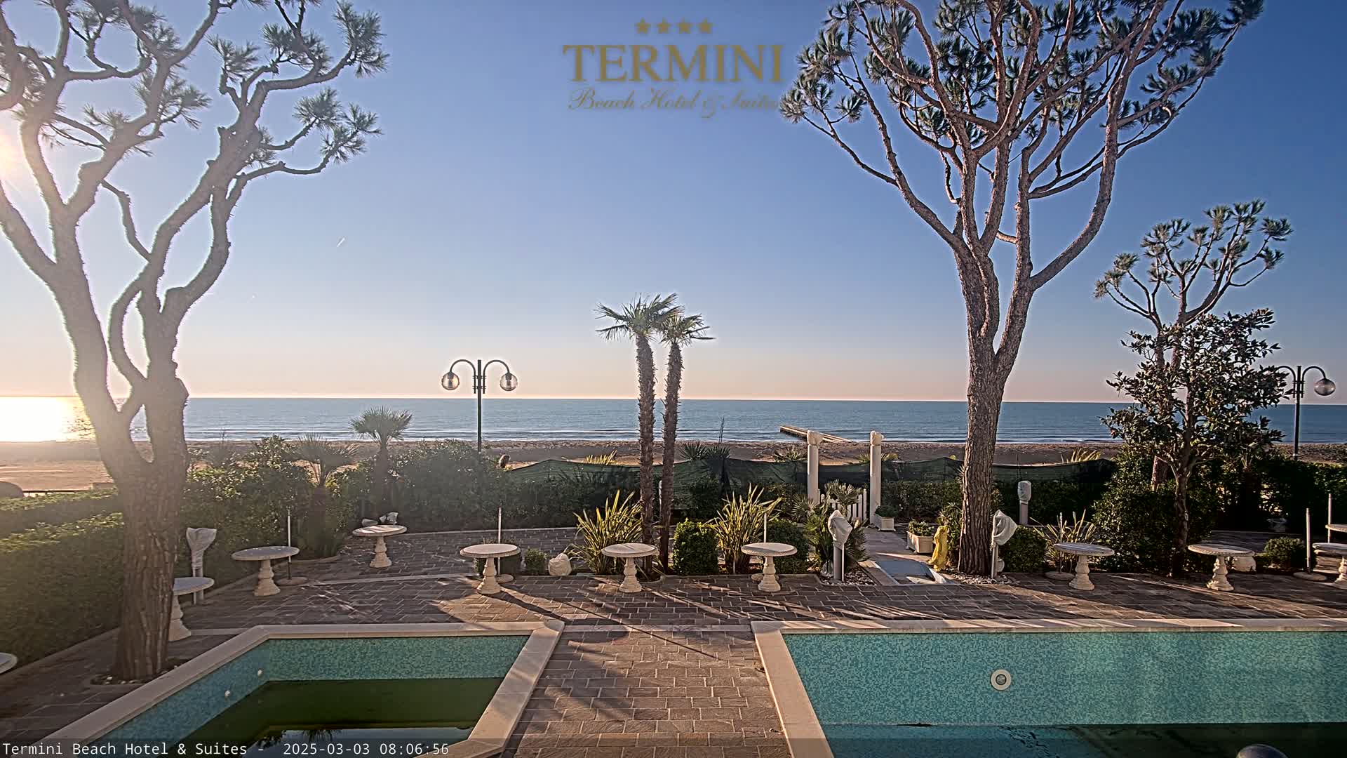 A tranquil beachfront patio with two swimming pools overlooks a calm ocean under a clear, sunny sky.
