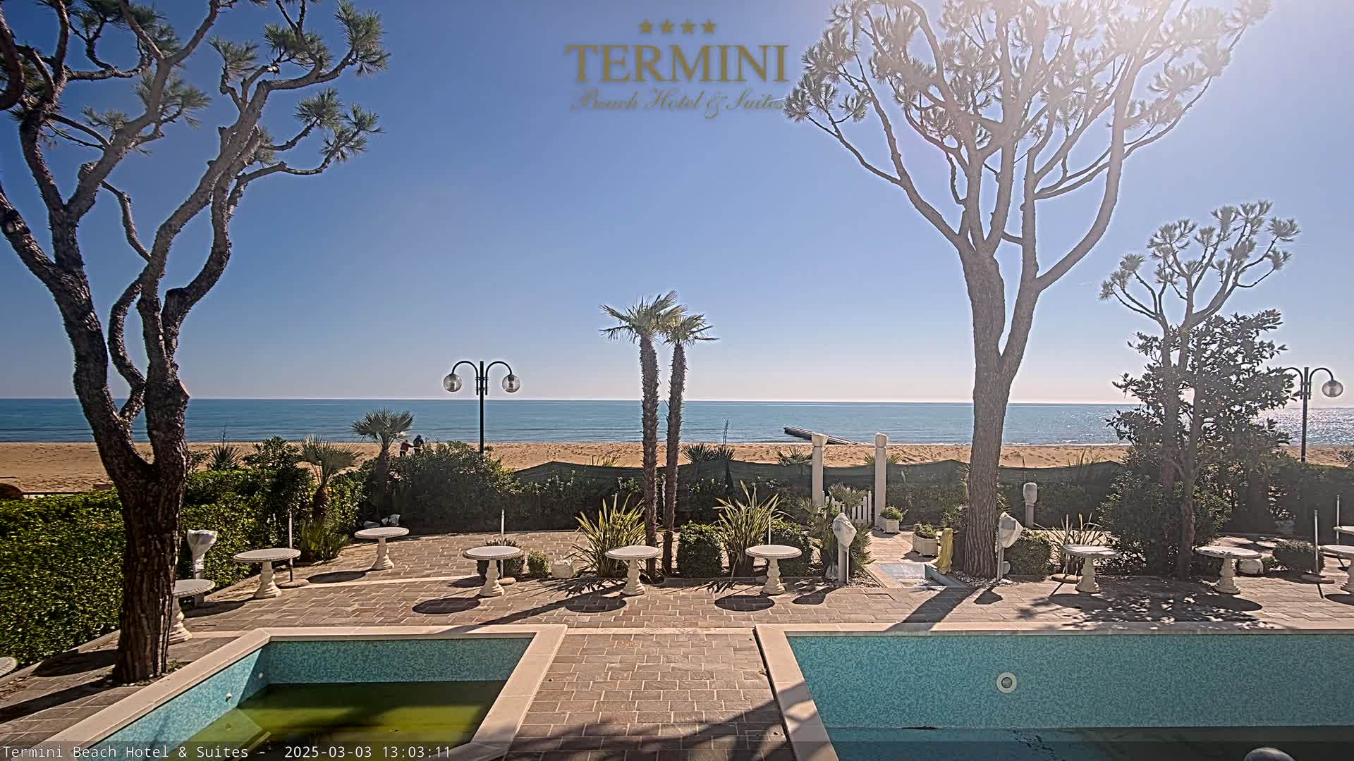 A sunny outdoor patio with a swimming pool overlooks a calm ocean beach.