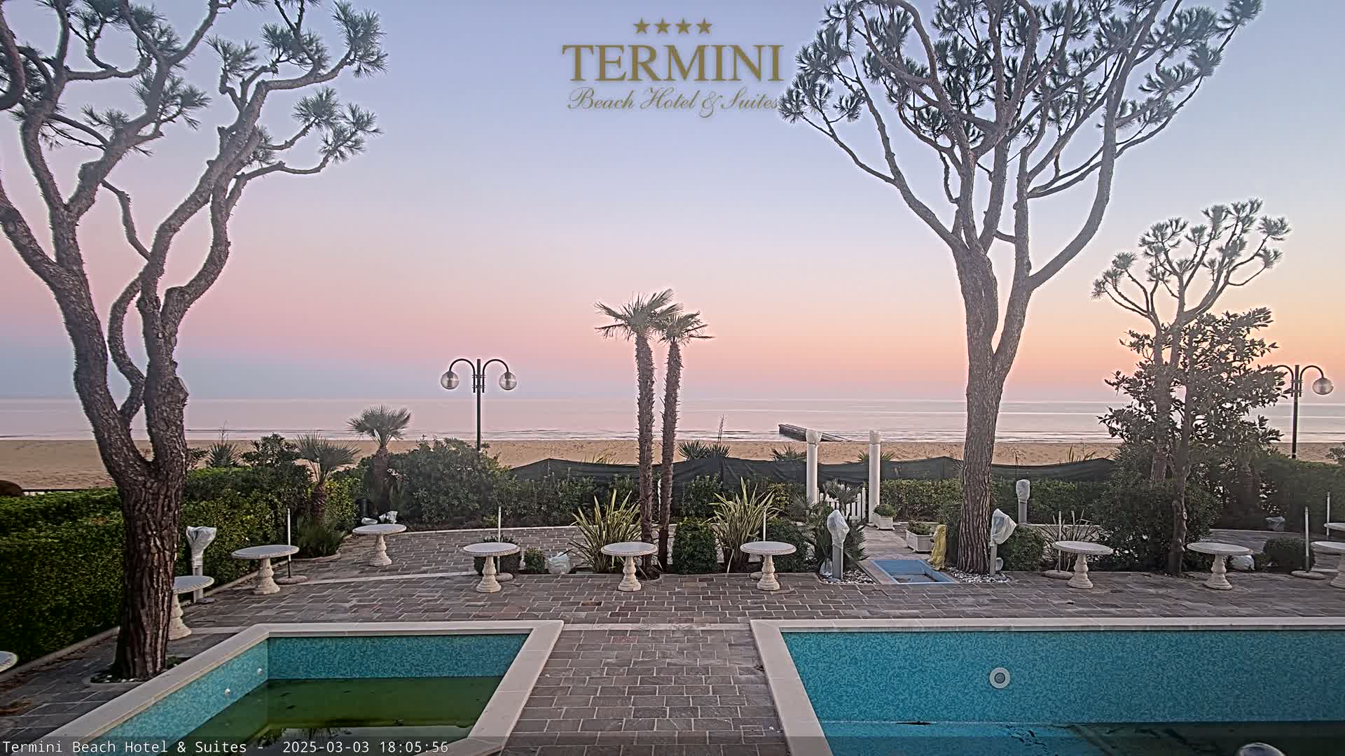 A tranquil outdoor patio scene at sunset overlooks a calm ocean beach, featuring two swimming pools and several small tables nestled among trees and landscaping.
