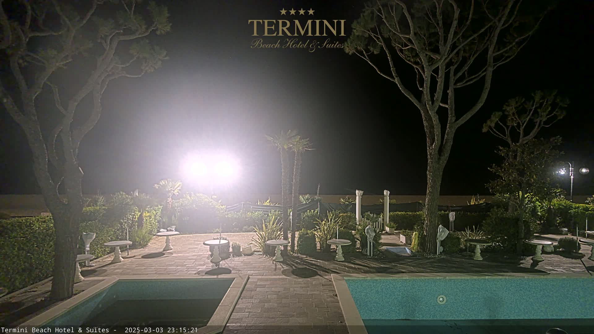 A nighttime view of a hotel patio area with a pool, several small tables, and palm trees, illuminated by bright lights.
