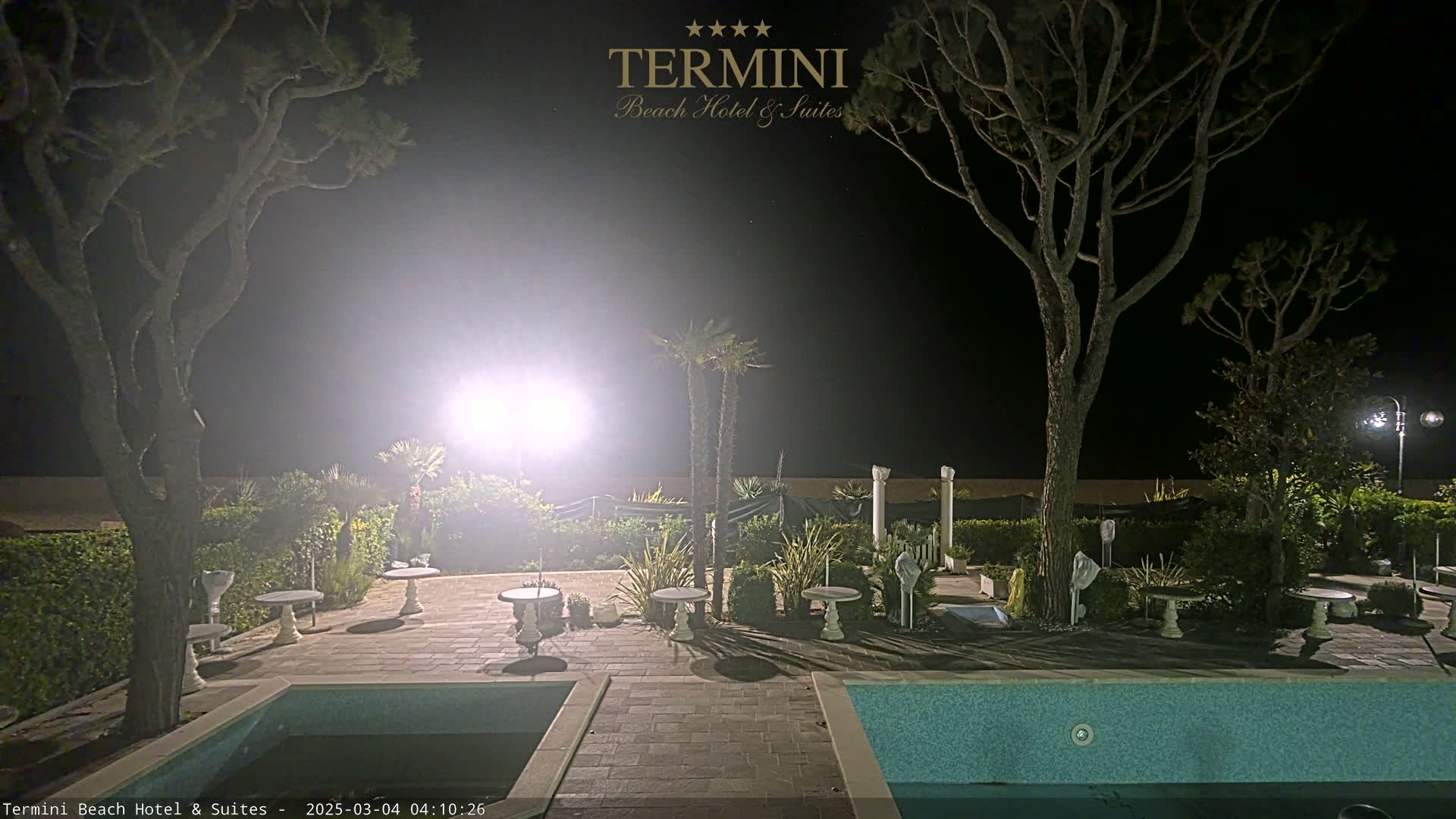 A nighttime view of a patio area with a swimming pool, illuminated by bright lights, surrounded by trees and landscaping.