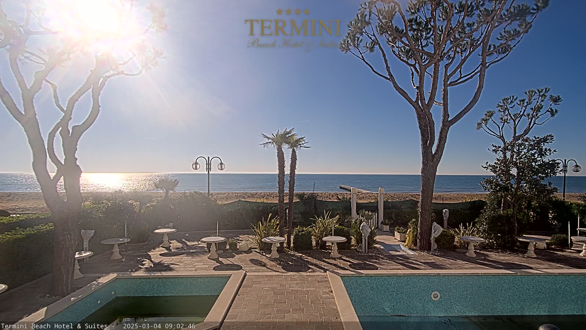 A sunny day at a beachfront hotel shows a patio with small tables and a partially visible swimming pool, overlooking a calm ocean and sandy beach.