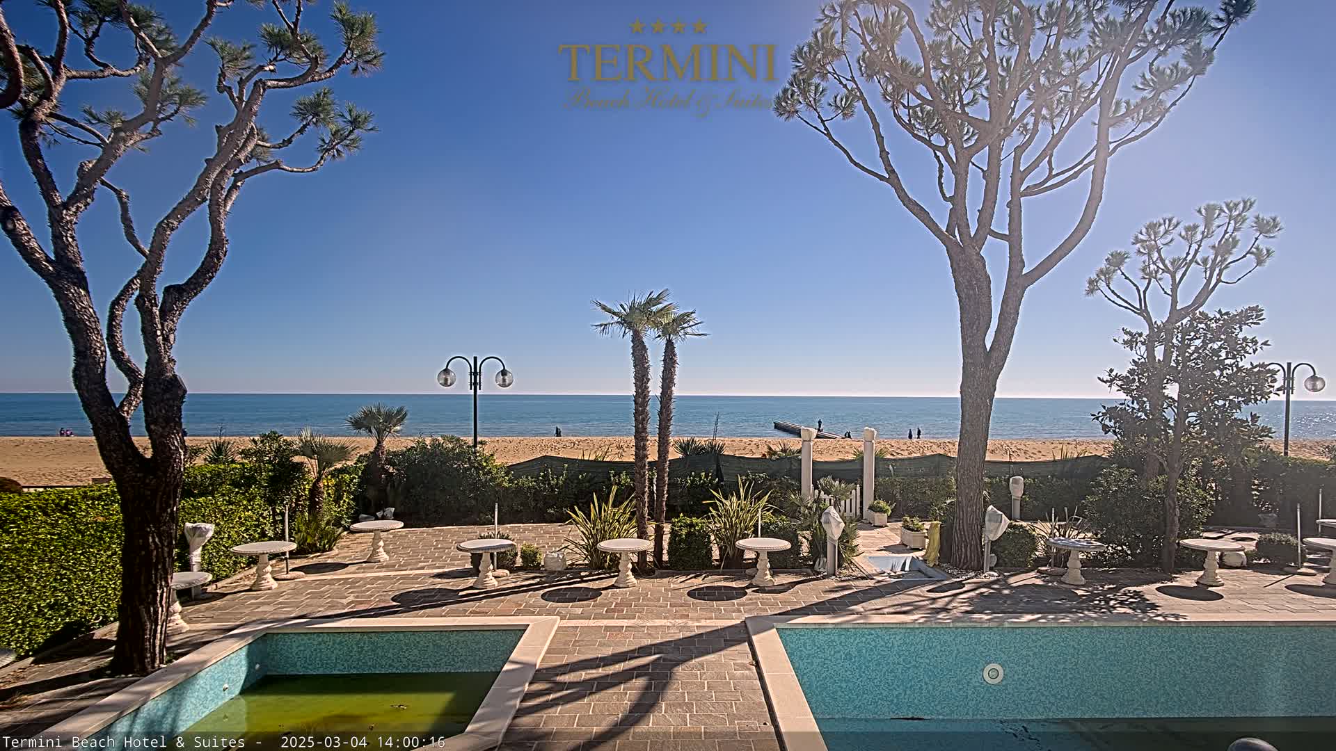 A sunny outdoor scene shows a patio area with two small pools, several small tables, and various shrubs and trees, all overlooking a sandy beach and calm ocean.