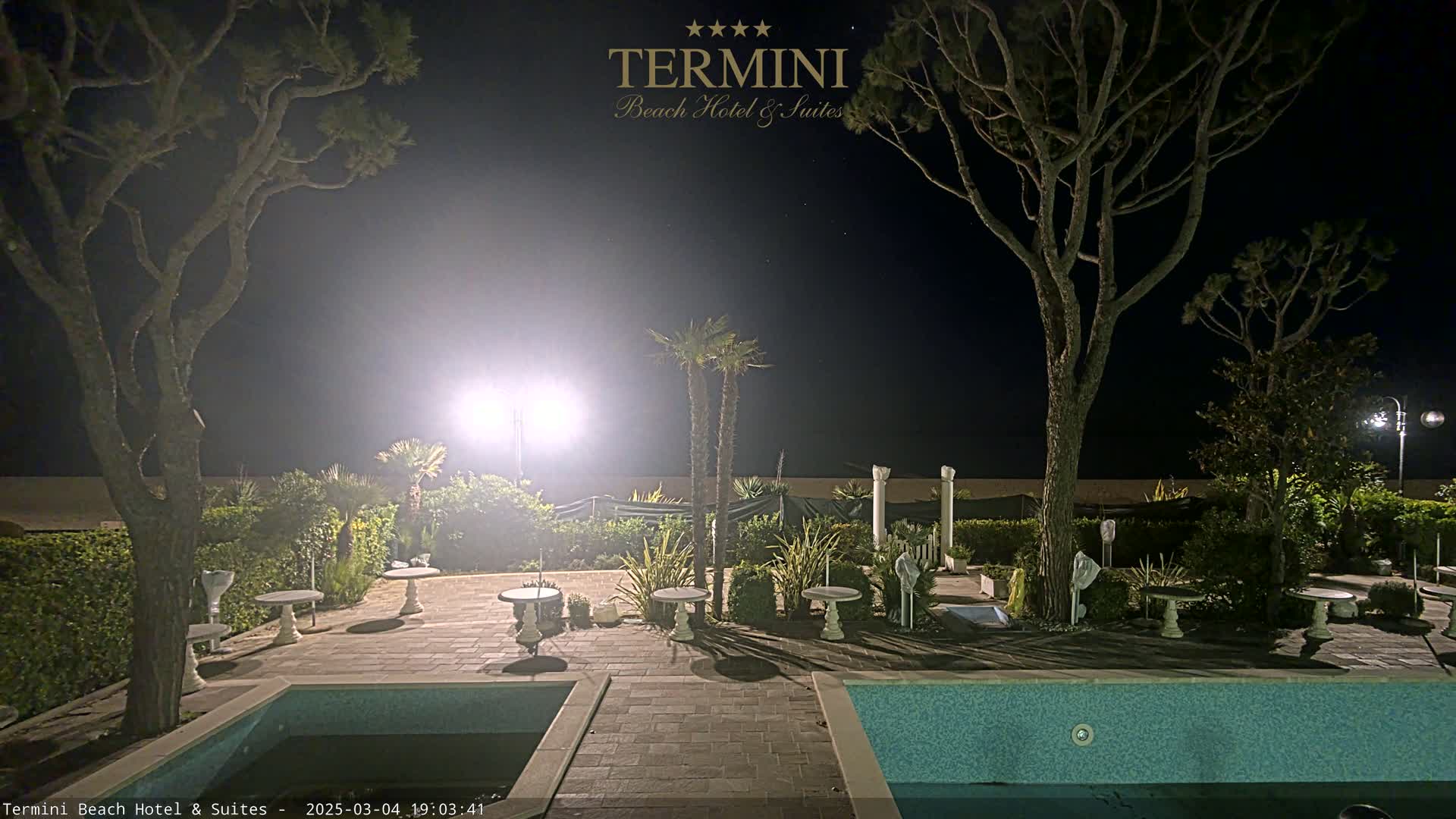 A nighttime view of a patio area with two swimming pools, several small tables, and palm trees, illuminated by bright lights, adjacent to a beach under a clear, dark sky.