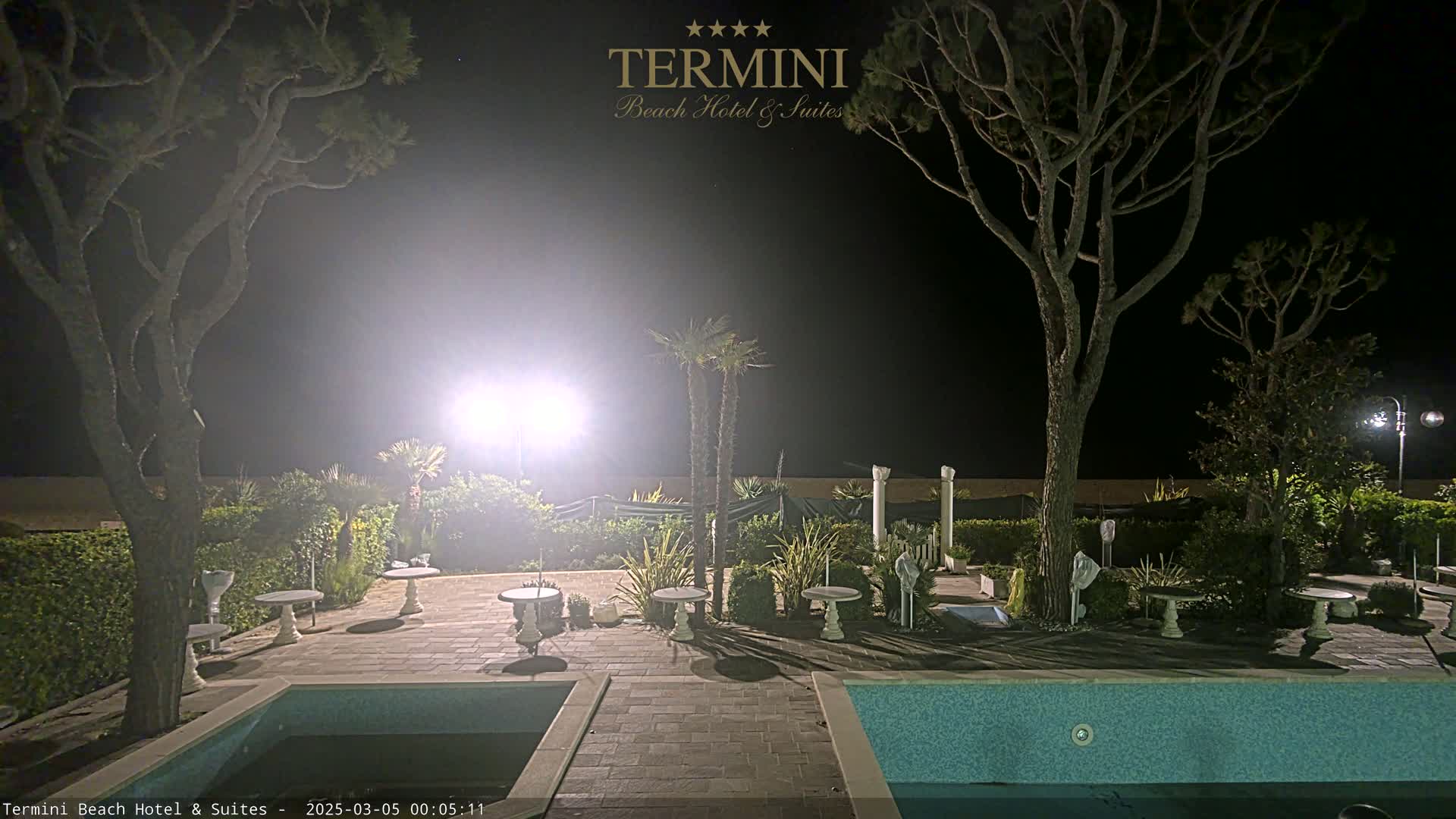 A nighttime view of a resort pool area with several small tables and trees surrounding it, illuminated by bright lights.