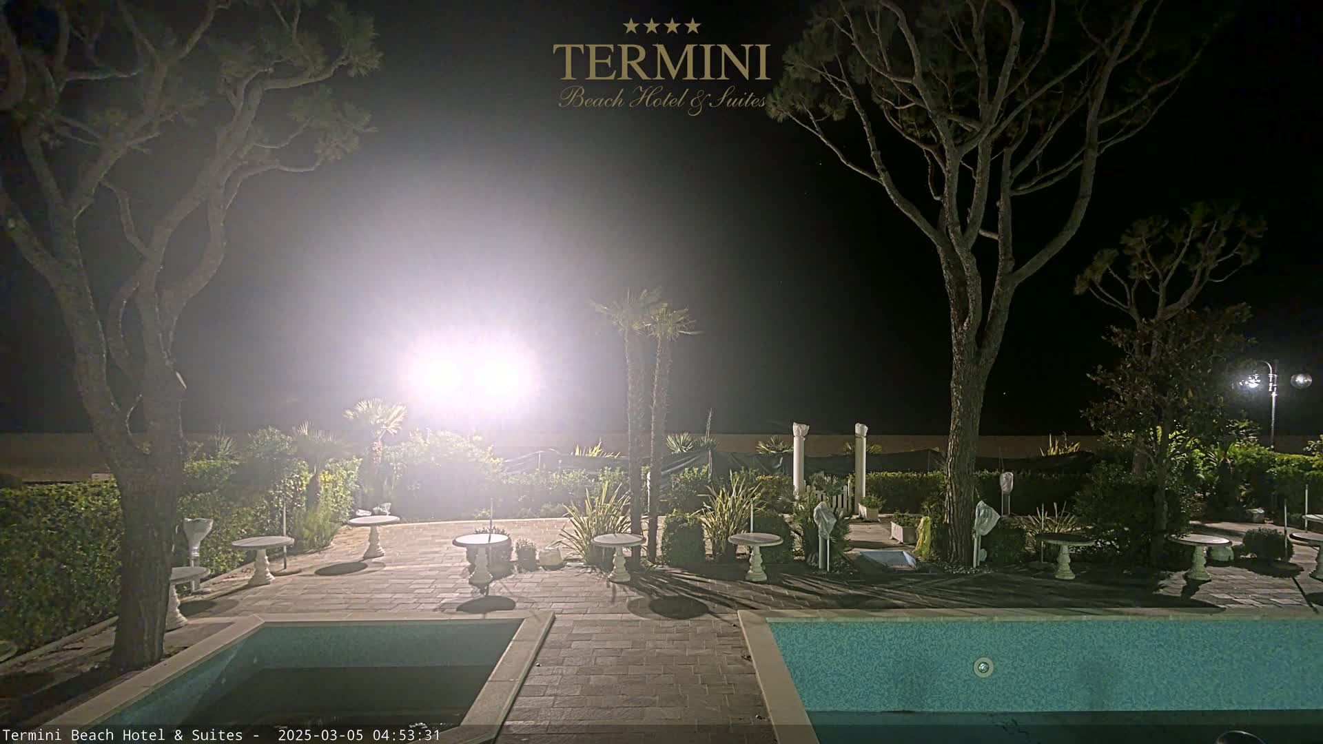 A nighttime view of a resort pool area with surrounding landscaping, trees, and patio furniture, illuminated by bright lights.