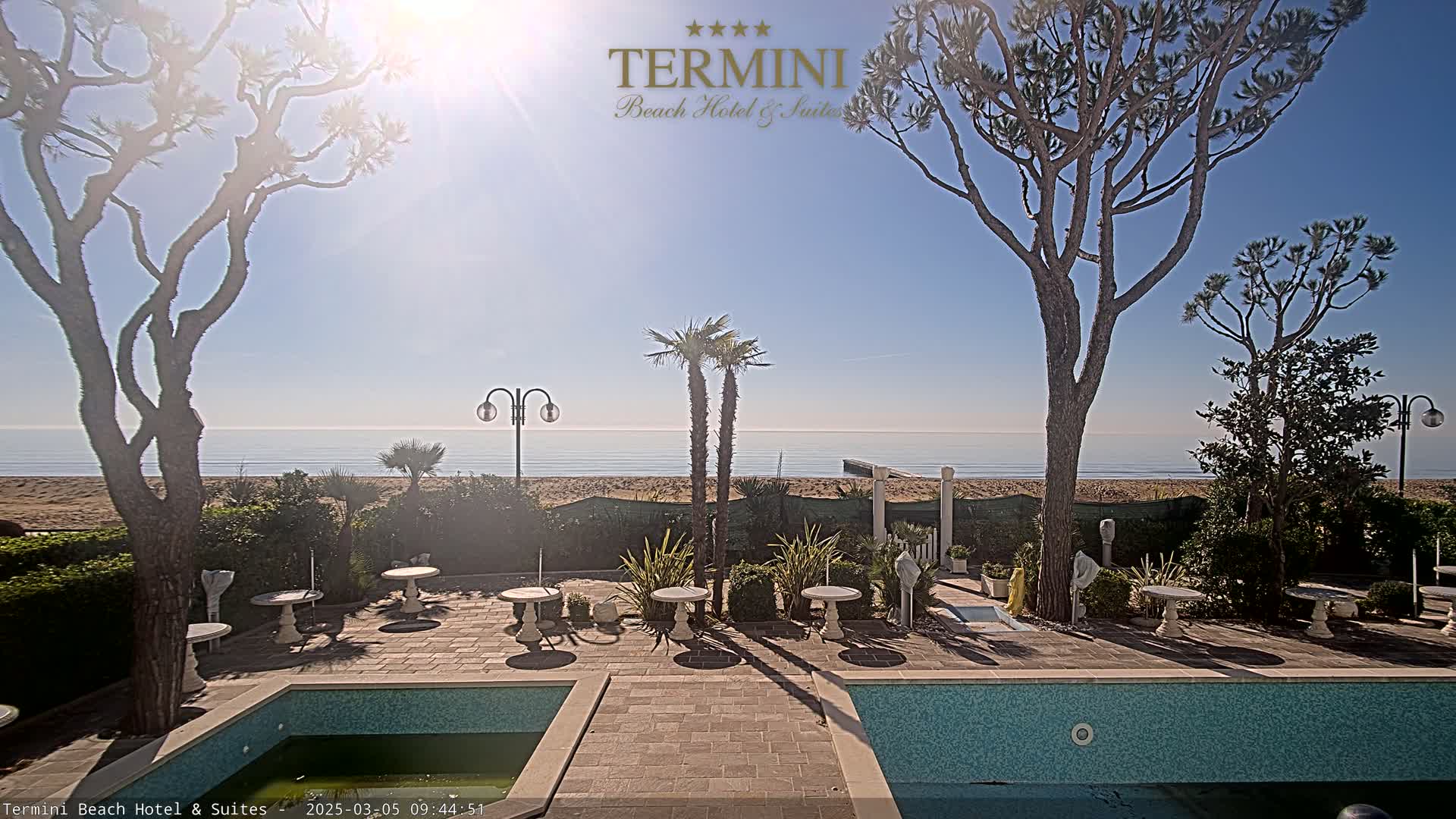 A sunny outdoor patio area with a swimming pool overlooks a sandy beach and calm ocean.