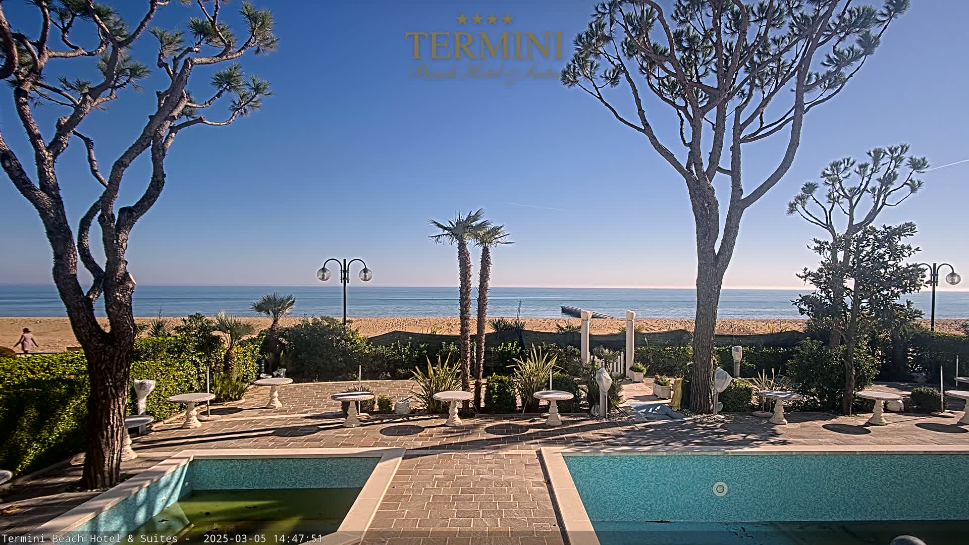 A patio area with two small pools overlooks a sandy beach and calm ocean under a clear blue sky.