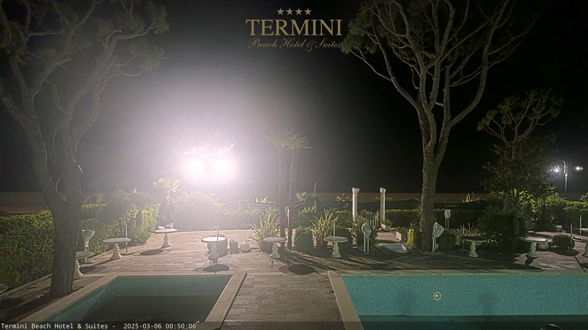 A nighttime view of a resort patio with a swimming pool, illuminated by bright lights, surrounded by trees and landscaping.