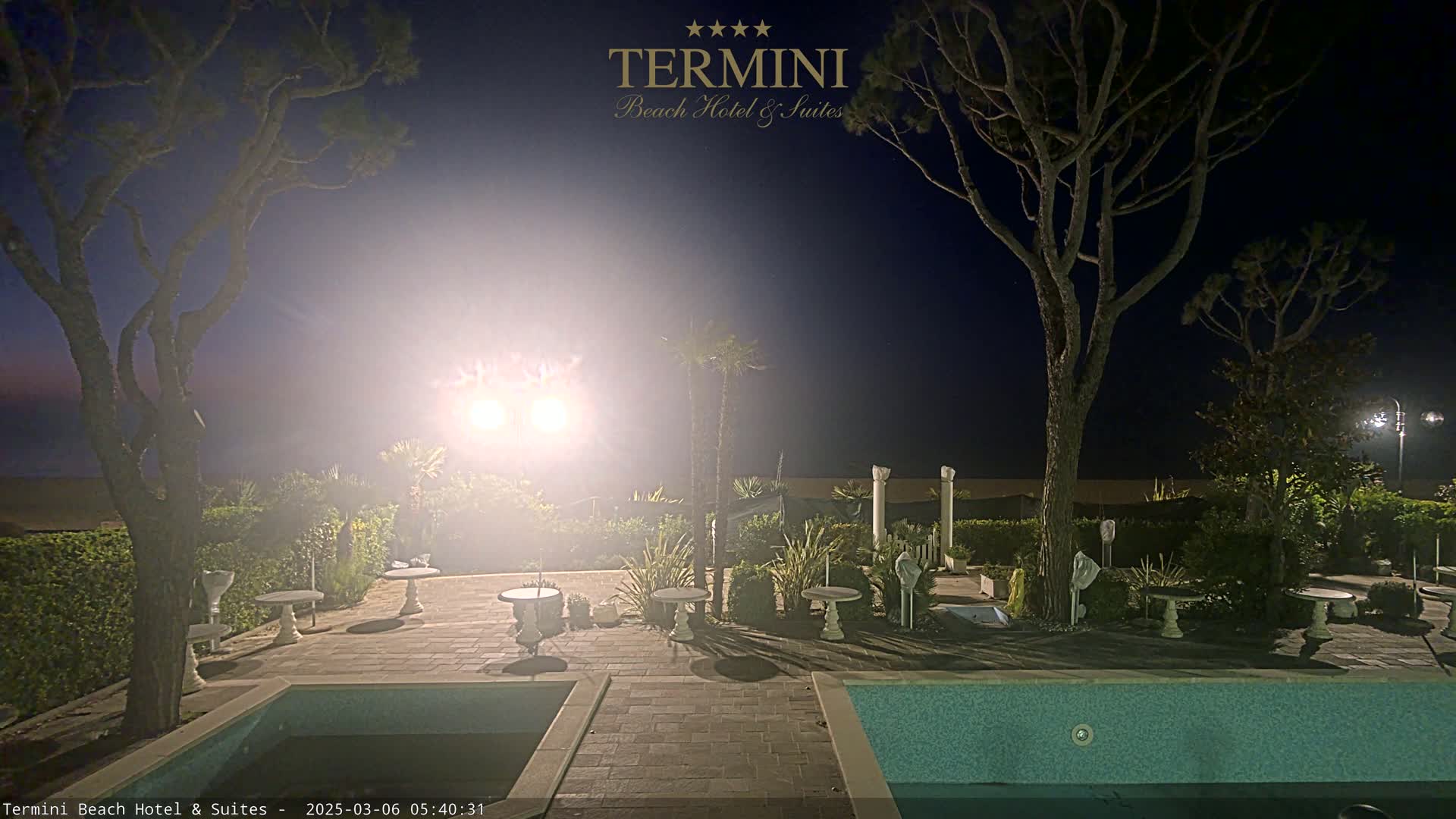 A nighttime view of a poolside patio area with several small tables and trees, illuminated by bright lights, under a dark sky.
