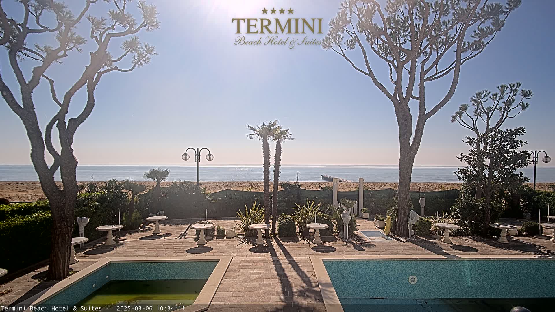 A sunny outdoor scene shows a patio area with two swimming pools, small tables and chairs, and several trees, all overlooking a sandy beach and calm ocean.