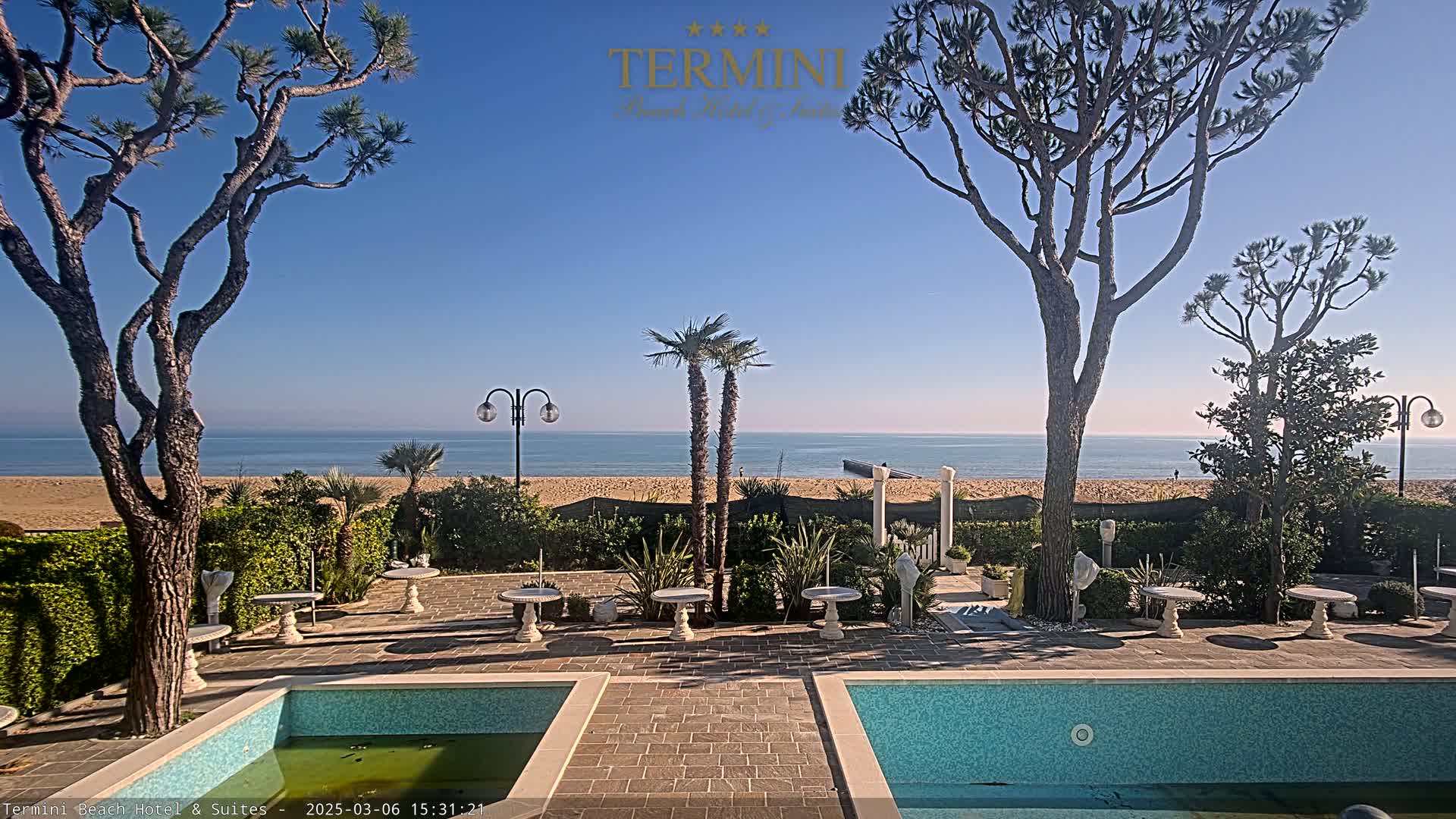 A clear, sunny day is shown overlooking a sandy beach and calm ocean from a patio with two small pools and landscaping featuring palm trees and stone tables.