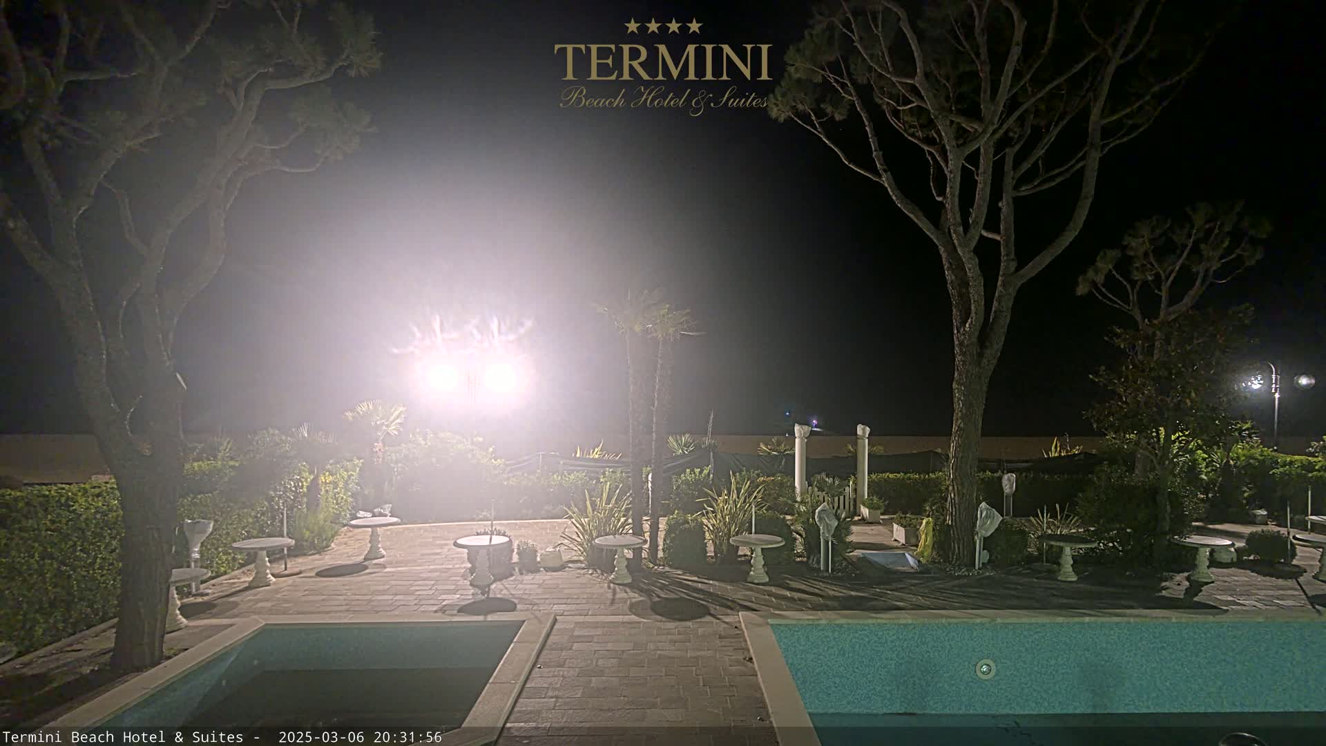 A nighttime view of a pool area with patio furniture and trees under hazy, bright lighting.