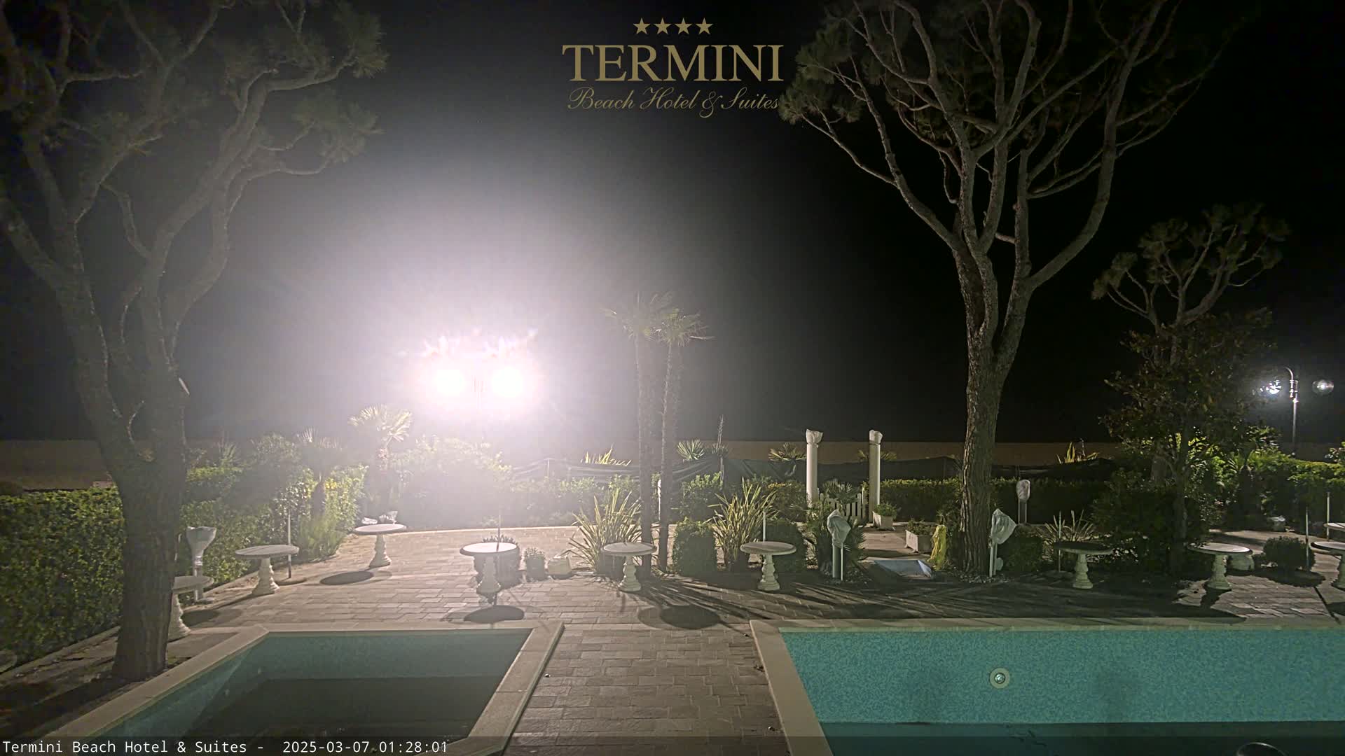 A nighttime view of a swimming pool area with surrounding landscaping and trees, illuminated by bright lights.