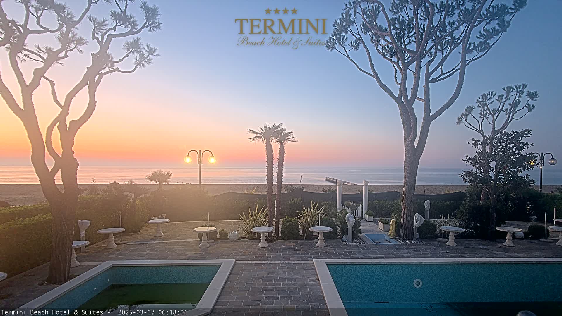 A tranquil sunrise over a calm ocean is visible from a patio area with two swimming pools, several small tables, and various trees and plants.