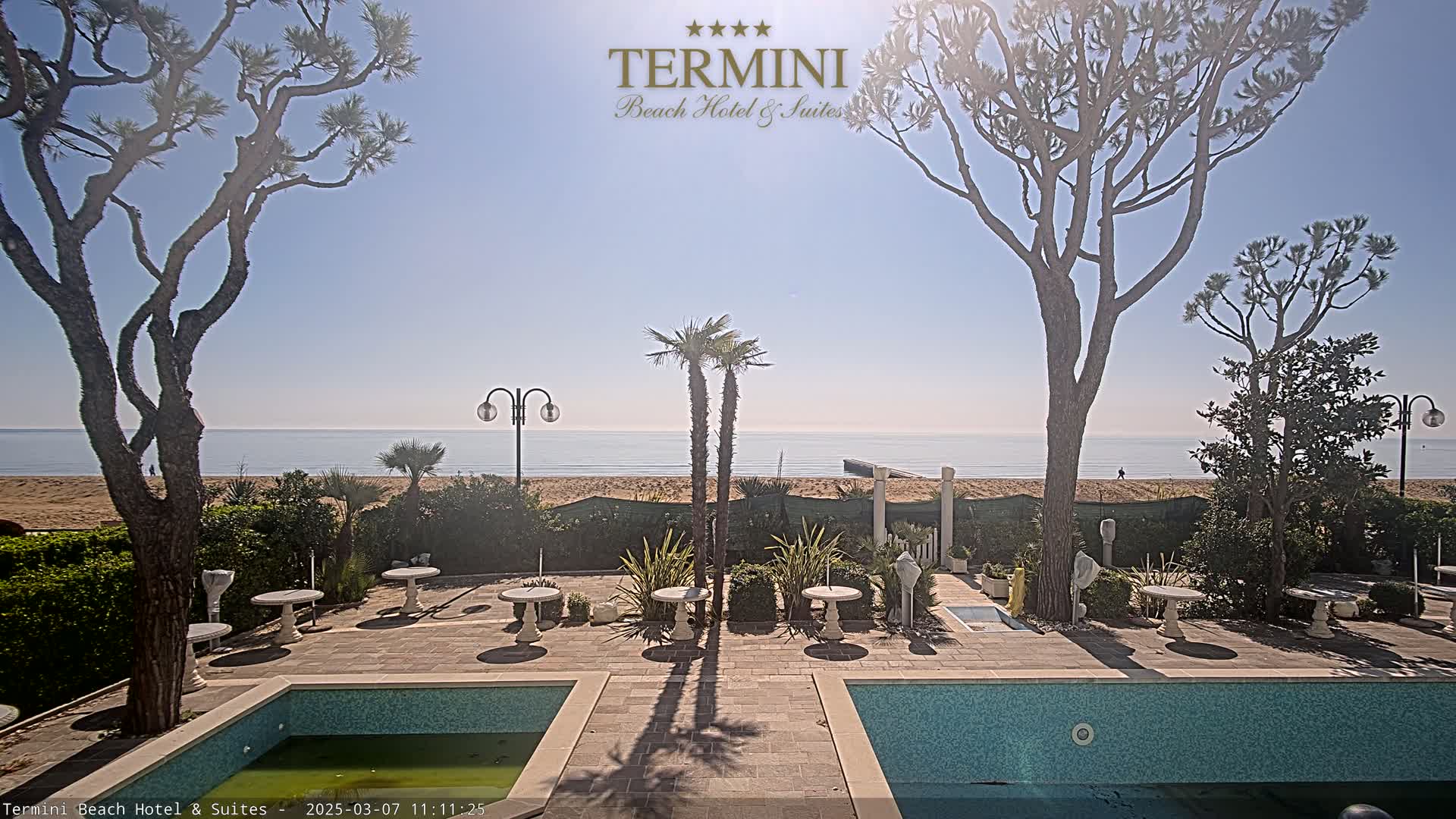 A sunny outdoor scene shows a patio area with two swimming pools, several small tables, and various trees, all overlooking a sandy beach and calm ocean.