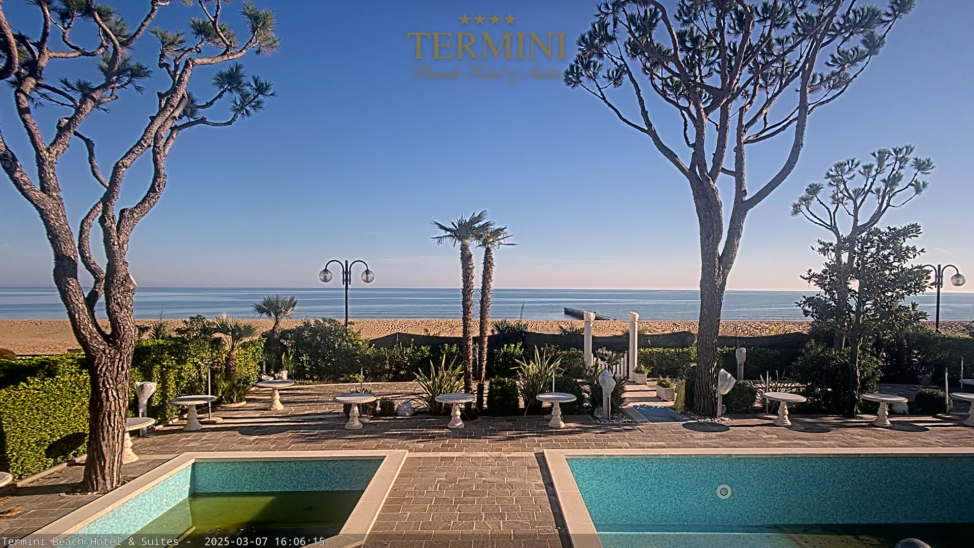 A patio area with two small pools overlooks a calm ocean and sandy beach under a clear blue sky.