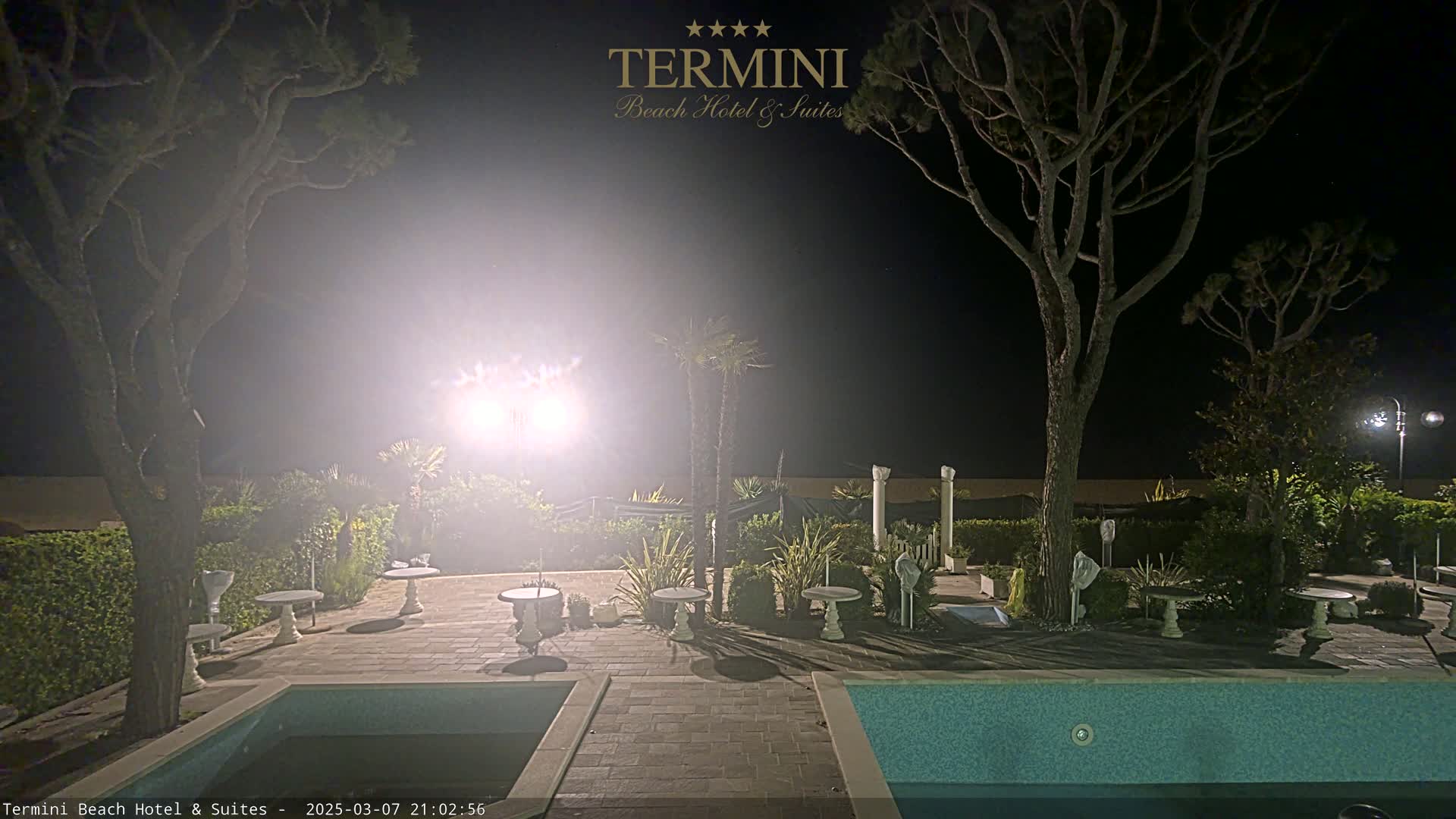 A nighttime view of a swimming pool area with patio furniture, trees, and bright lights illuminating the scene.