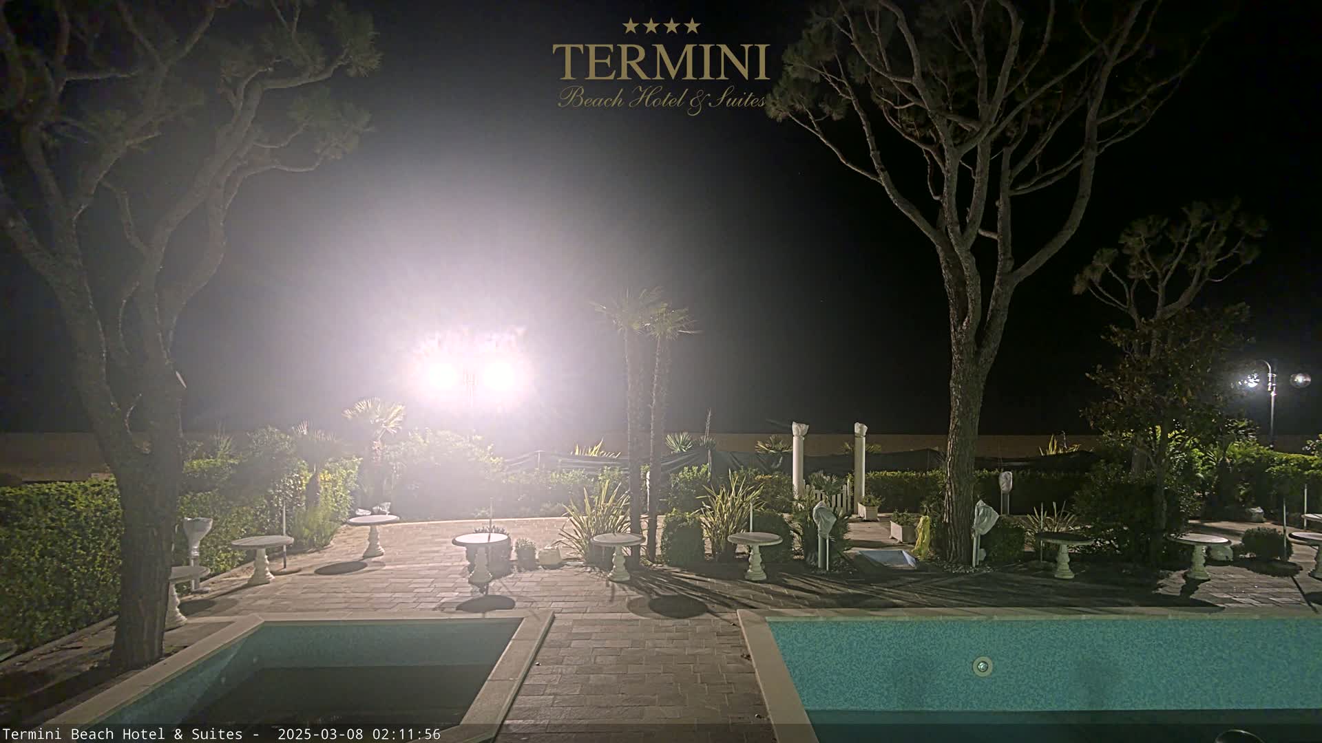 A nighttime view of a resort pool area with surrounding trees and small tables, lit by bright lights.